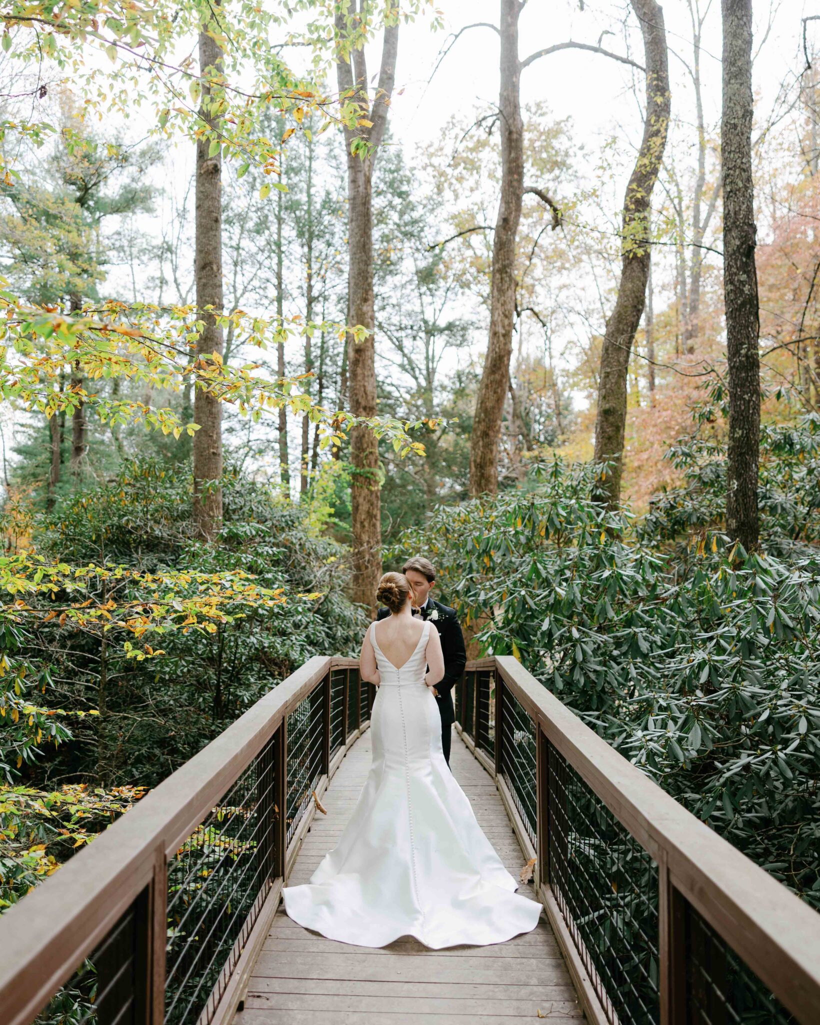 asheville wedding photographer A bride and groom stand facing each other on a wooden bridge in a lush forest, surrounded by tall trees and greenery, beautifully captured by an Asheville wedding photographer. The bride wears a white gown and the groom is in a dark suit.