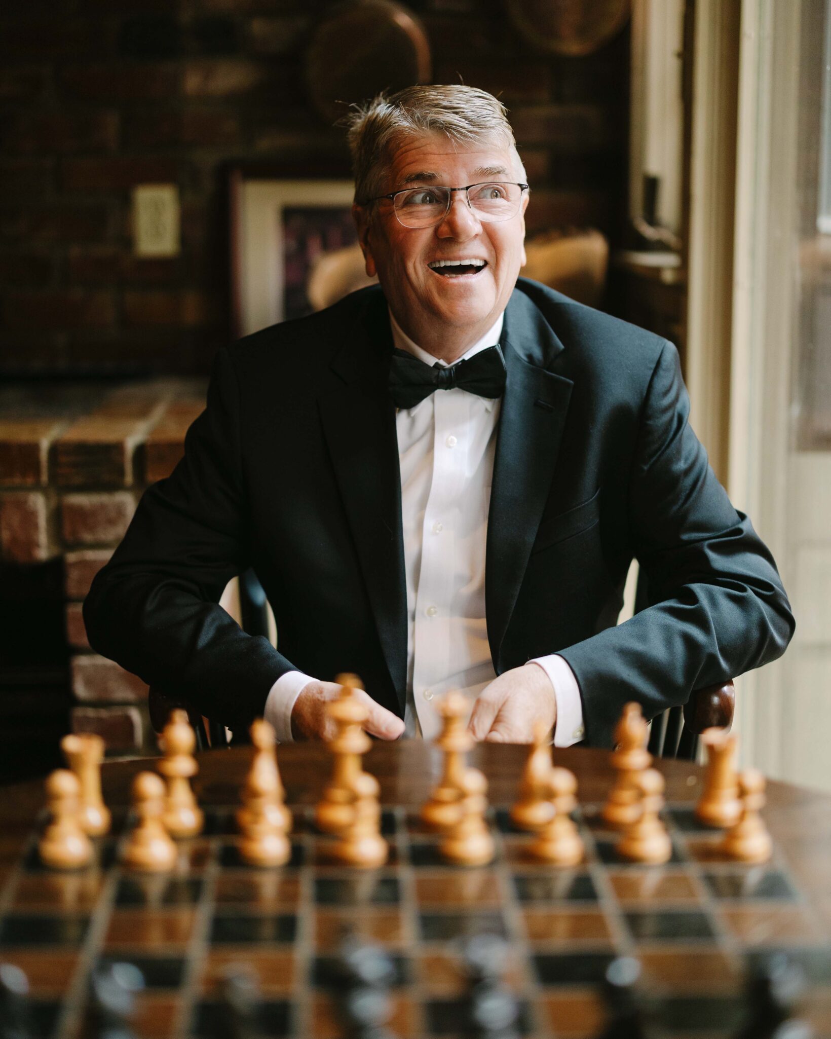 asheville wedding photographer A smiling man in a tuxedo sits by a window with a chessboard in front of him, appearing happy and engaged in a cozy room with brick walls—captured by an Asheville wedding photographer.