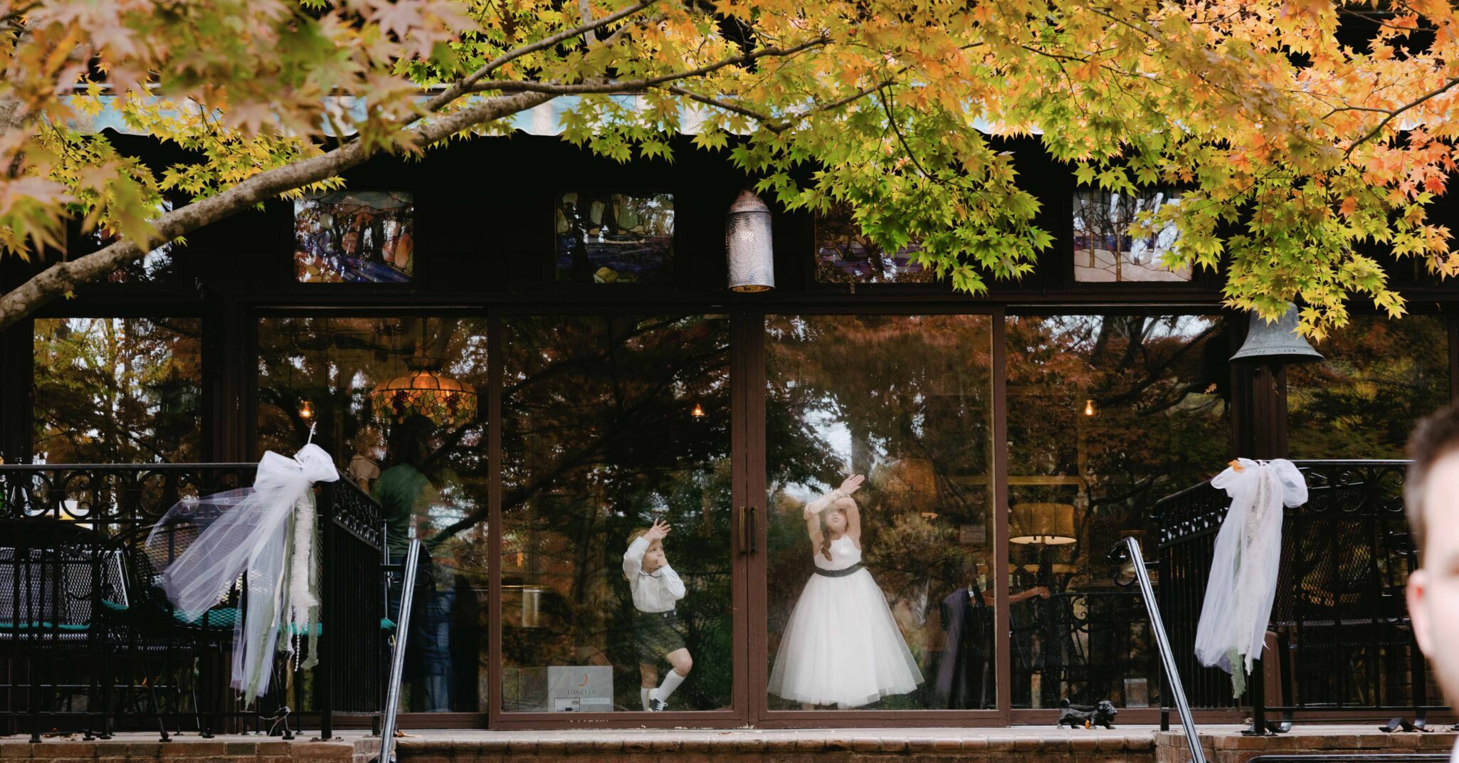 asheville wedding photographer Two children, one in a white dress and one in a white shirt and shorts, stand behind large glass doors adorned with white ribbons, framed by autumn leaves—captured perfectly by an Asheville wedding photographer.