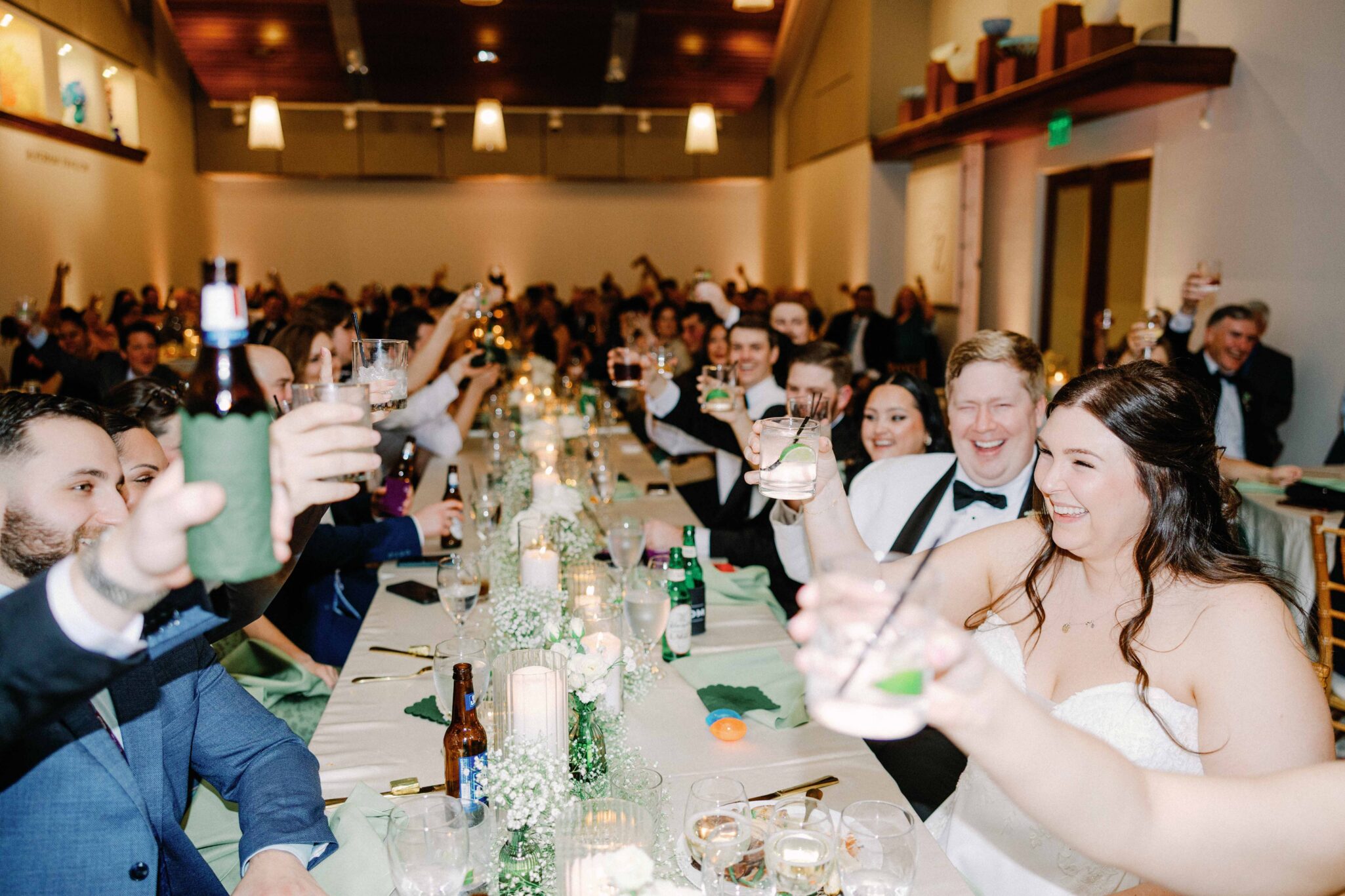 asheville wedding photographer A large group of people dressed formally sit at a banquet table, raising glasses and smiling in celebration. The room, captured by an Asheville wedding photographer, is brightly lit and decorated for a festive wedding reception.