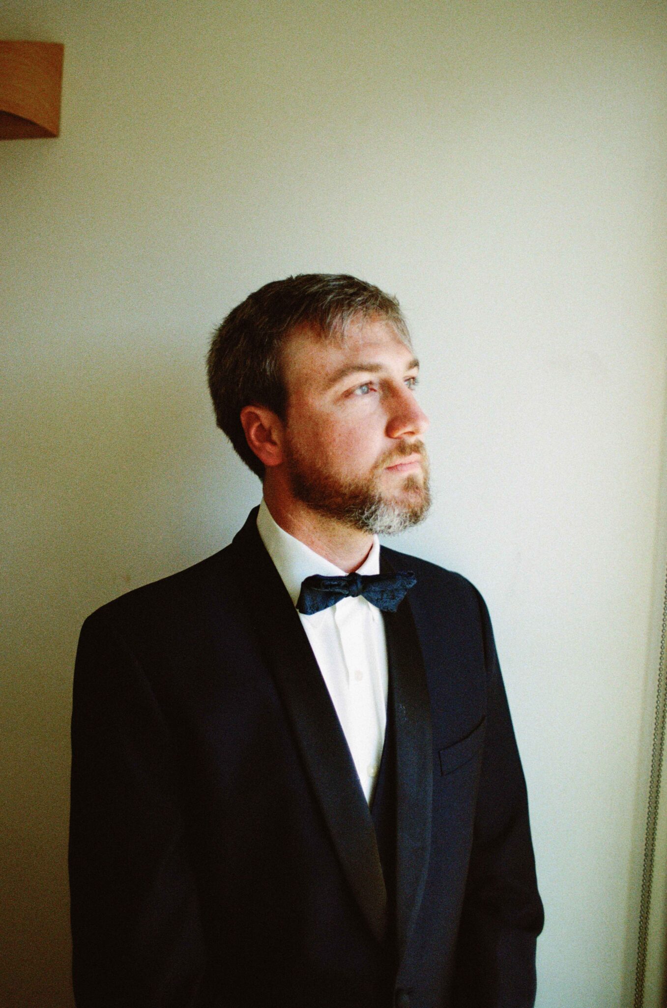 A man with short brown hair and a beard, wearing a black tuxedo with a bow tie, stands indoors looking thoughtfully to the side. The background is a plain, light-colored wall.