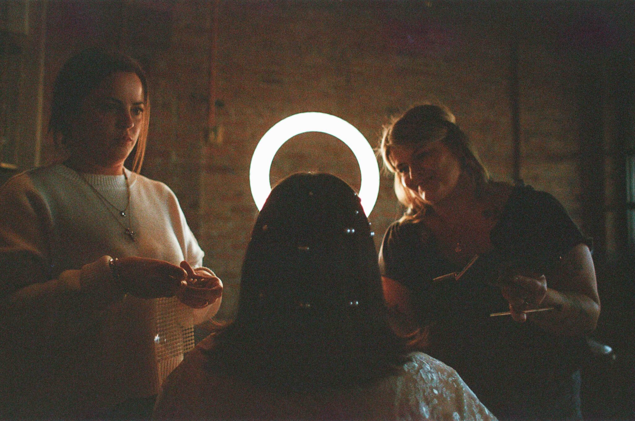 Two women stand on either side of a seated person whose back faces the camera, with a bright ring light behind them creating a halo effect. The women appear to be styling the seated persons hair.
