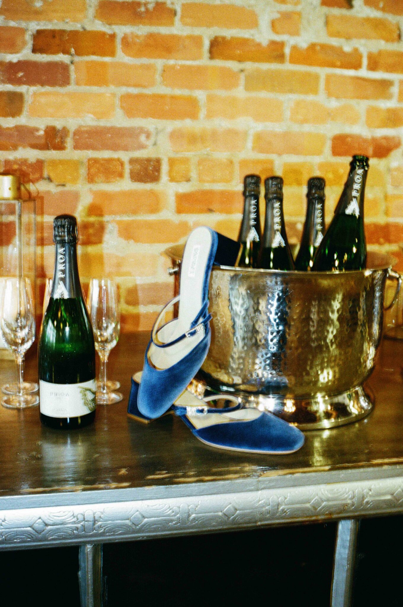 Bottles of sparkling wine chill in an ice bucket on a table set with wine glasses, while blue velvet shoes rest nearby—one propped on the bucket's edge. The brick wall hints at getting ready in a downtown Asheville venue, captured on 35mm film.