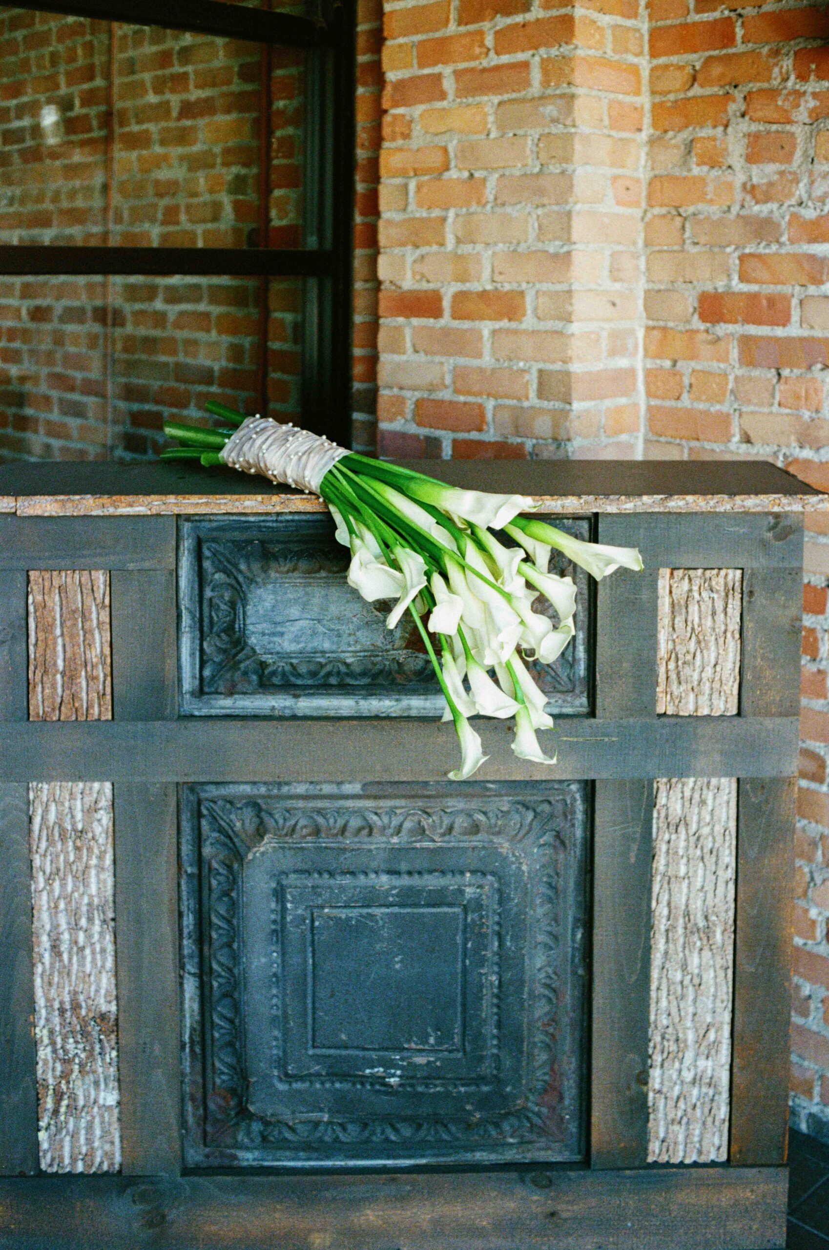 A bouquet of white calla lilies rests on a rustic, dark wooden table with bark details, set against a background of exposed brick walls and a window.