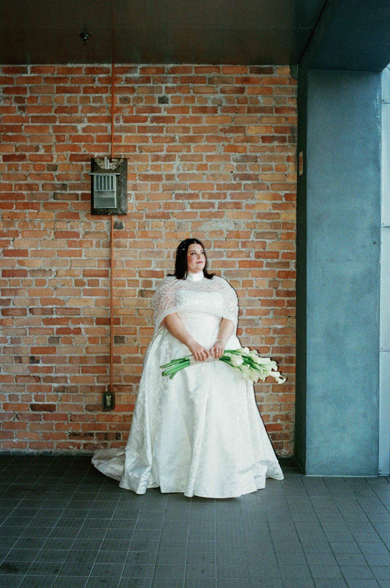 A bride in a white dress and shawl stands against a brick wall, holding a bouquet of white flowers and looking slightly to the side.