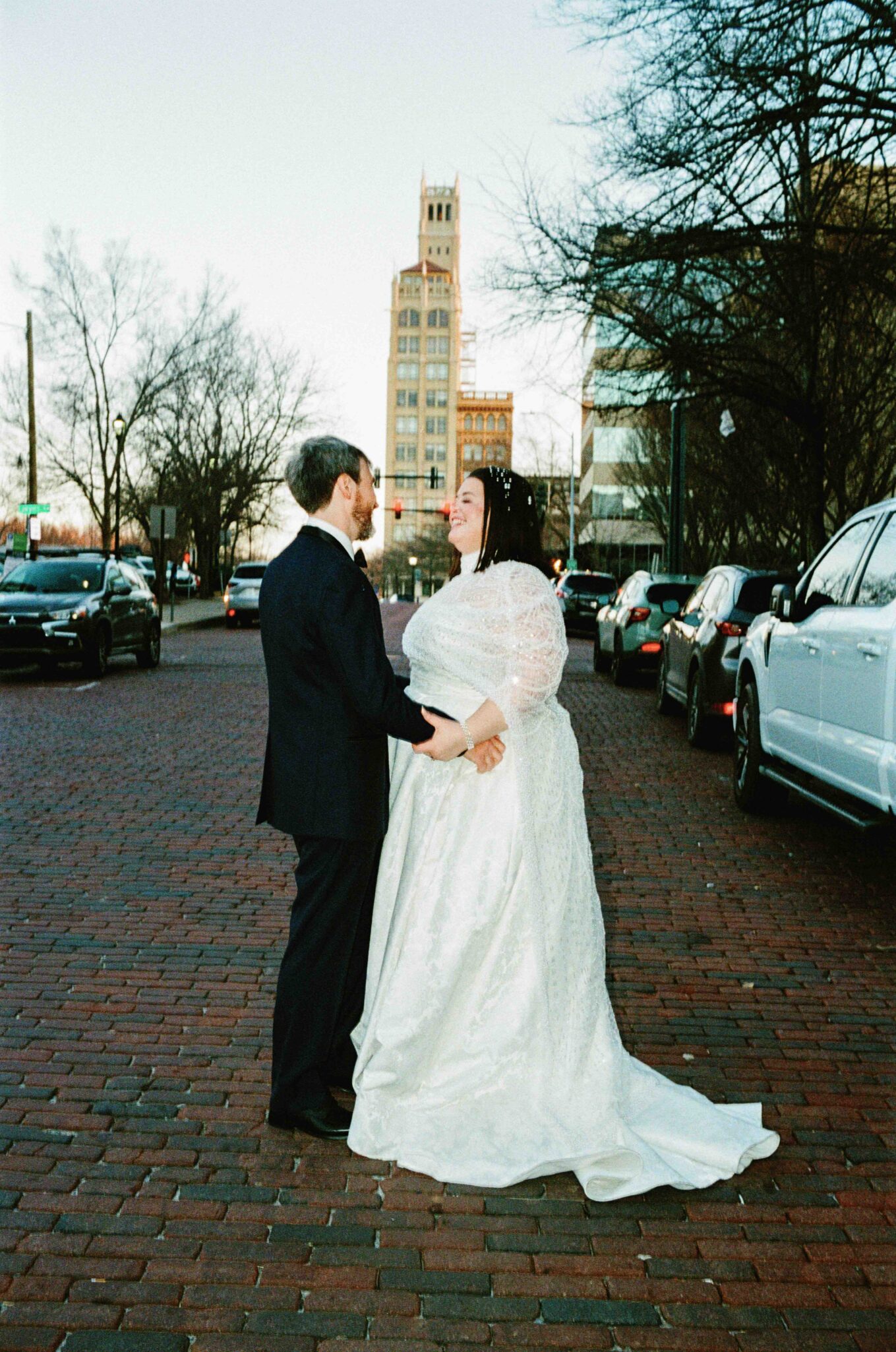 A bride in a white gown and a groom in a dark suit stand holding hands and smiling at each other on a brick street with cars parked along the sides and a tall building in the background.