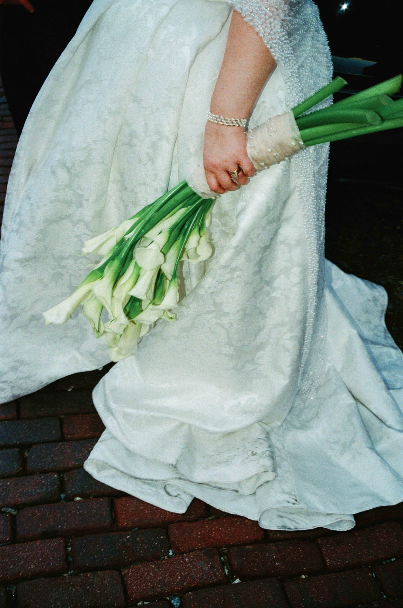 A bride in a white, textured gown holds a bouquet of white calla lilies, with her arm adorned by a sparkling bracelet, standing on a brick pathway.