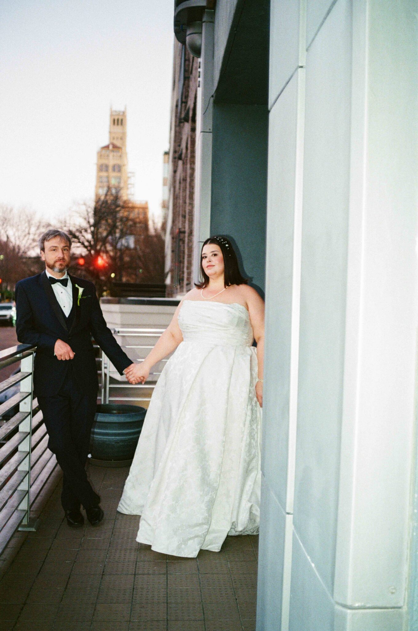A bride in a white strapless gown and a groom in a black tuxedo stand holding hands on an outdoor balcony. The bride leans against a wall, while the groom stands nearby. A tall building is visible in the background.
