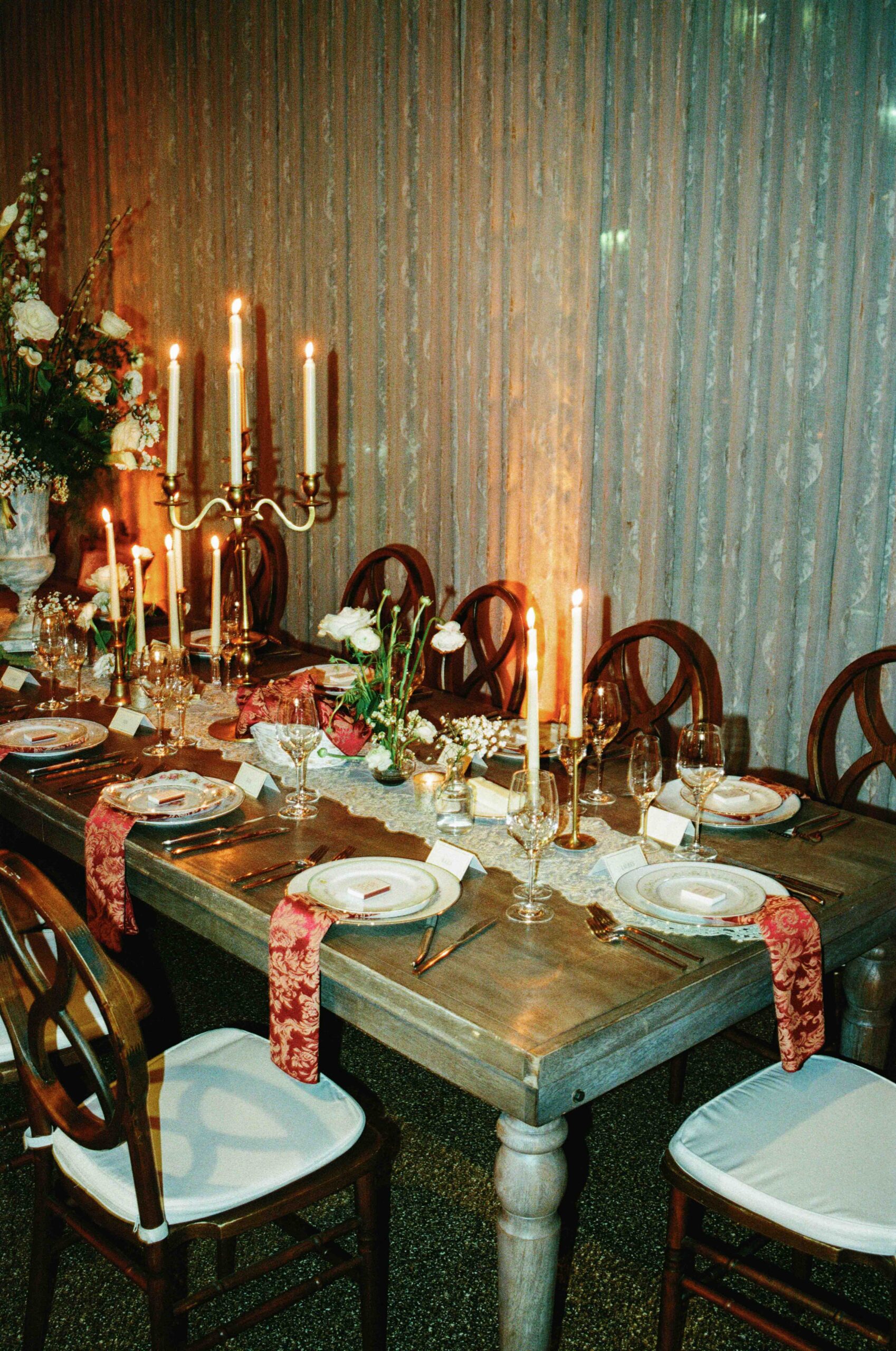 An elegant dining table set for a formal dinner, featuring lit candles, glassware, plates with red napkins, floral centerpieces, and wooden chairs with white cushions, against a backdrop of sheer curtains.