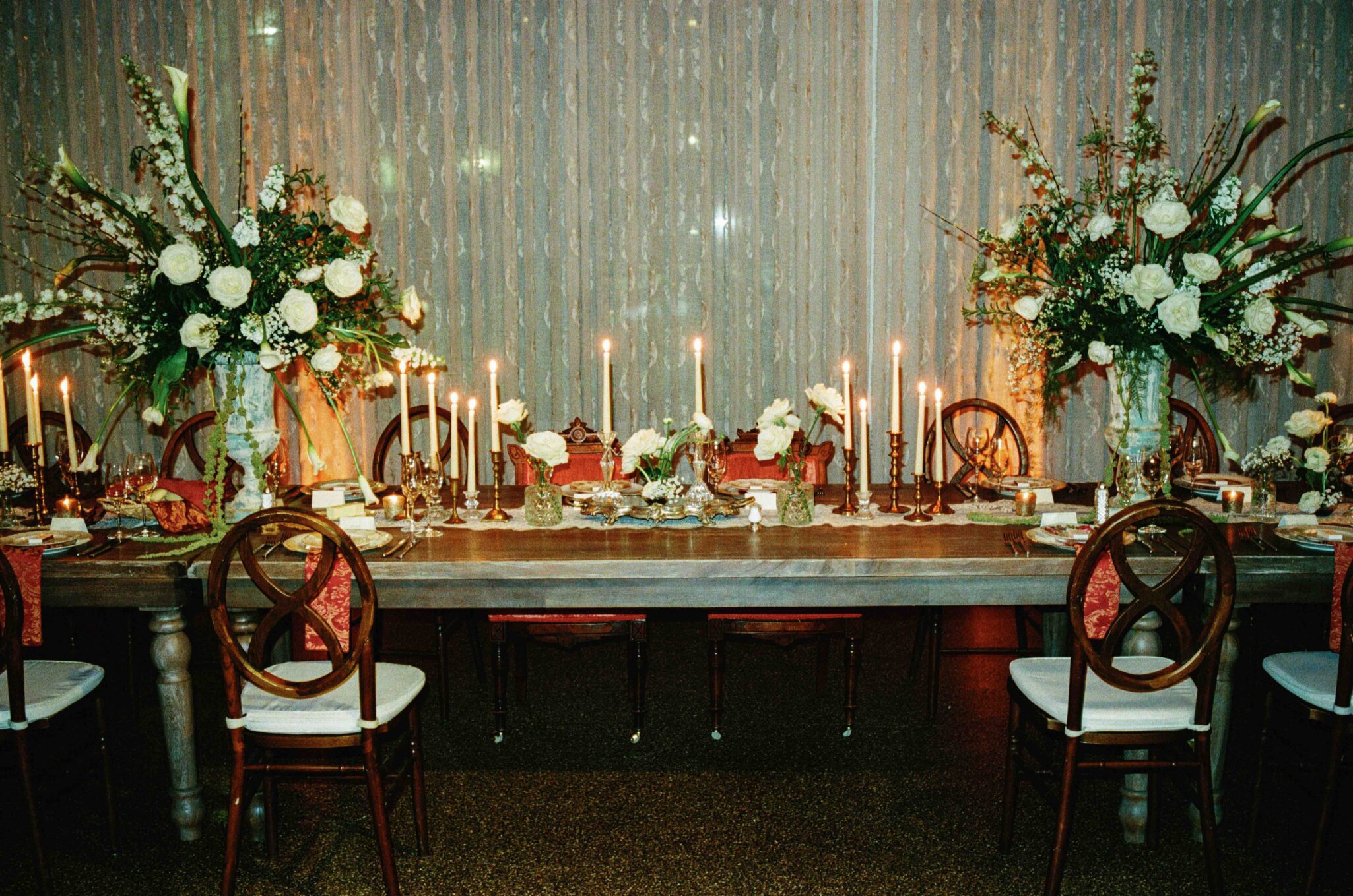 A formal dining table set with elegant place settings, tall floral arrangements, and lit candles. The chairs are wooden with white cushions, and a sheer curtain hangs in the background. The scene is warm and inviting.