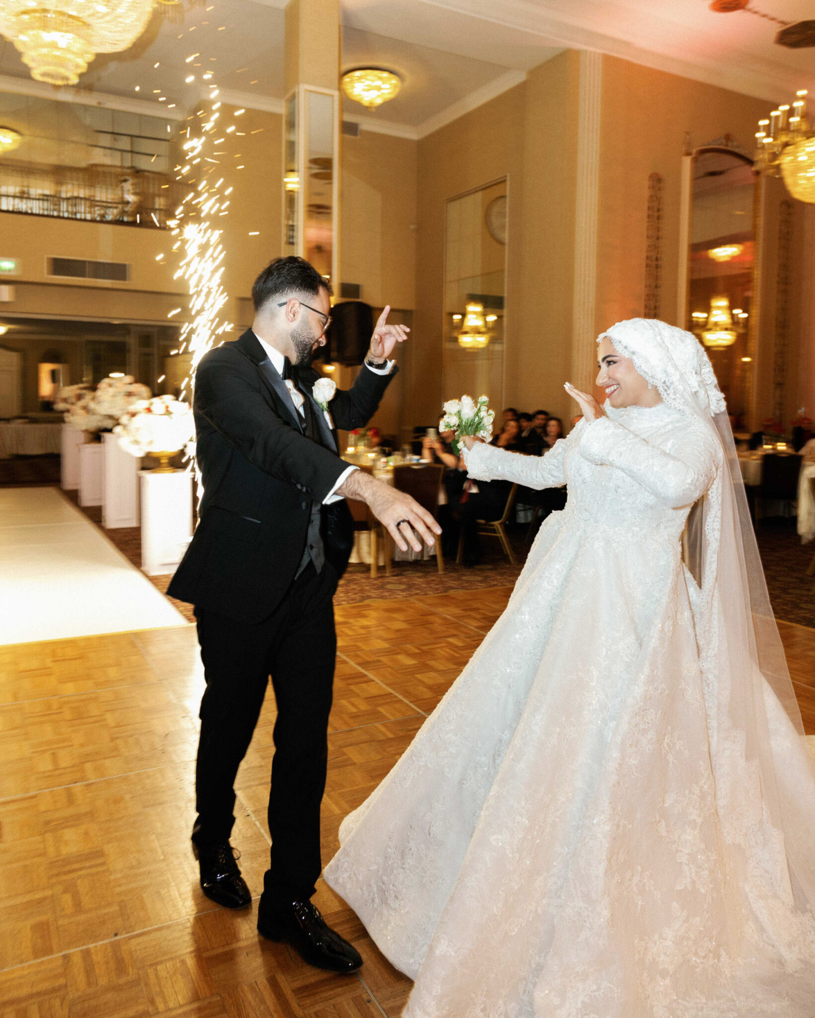 A bride in a white gown and hijab dances joyfully with a groom in a black suit on a wooden dance floor, as Chicago Arab wedding photography captures the white floral decor and bright indoor fireworks at their unforgettable reception.