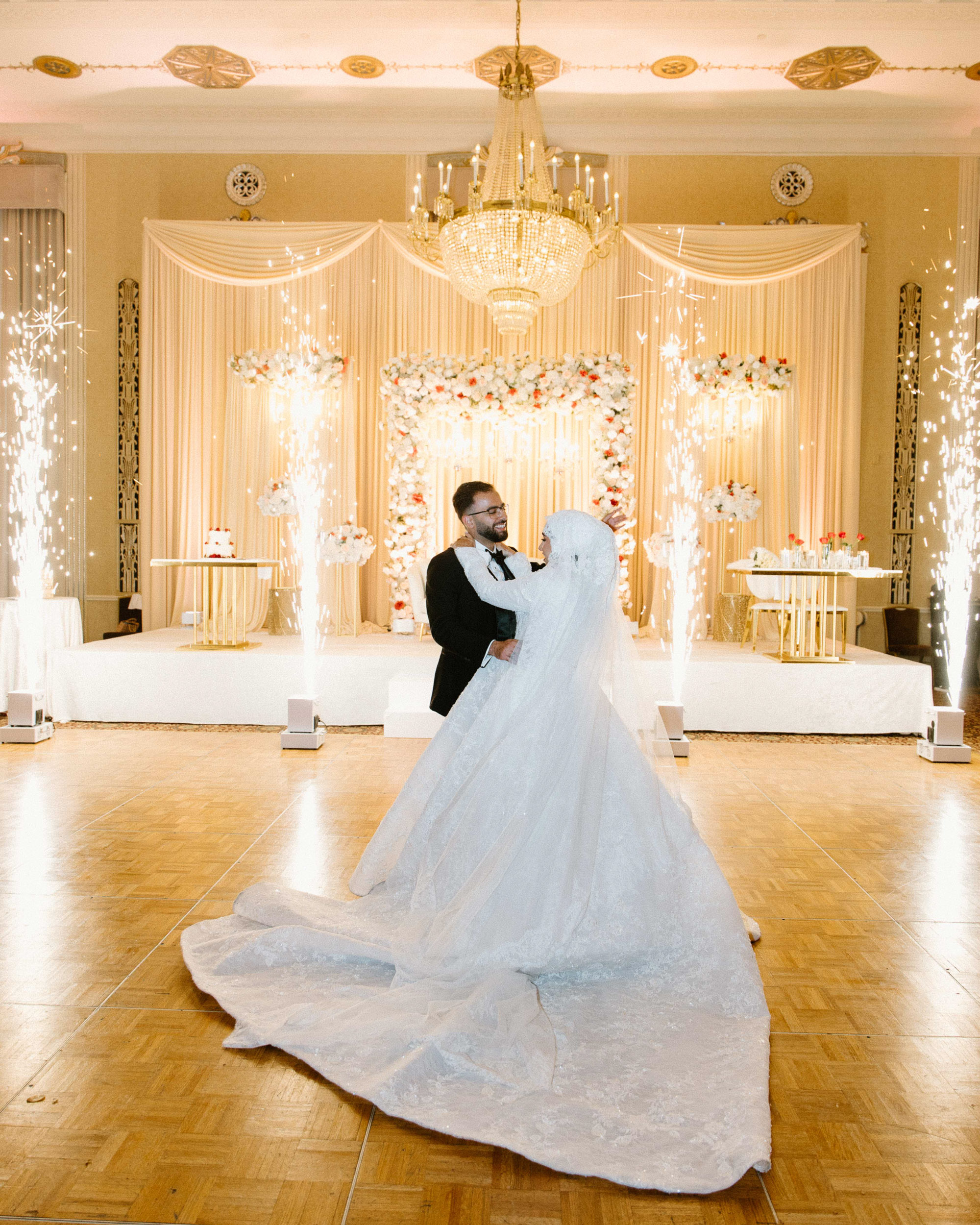 At an Arab wedding, the bride and groom share their first dance in an elegant ballroom, surrounded by sparklers and a floral arch. The chandelier above adds glamour to this multi cultural weddin celebration.