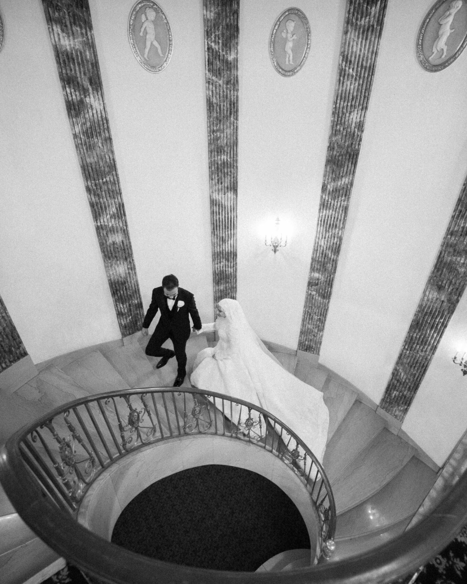 A bride in a long white gown and a groom in a black tuxedo walk down a curved staircase in an elegant, ornate setting, viewed from above—perfect for capturing the magic of Arab wedding photography in Chicago.
