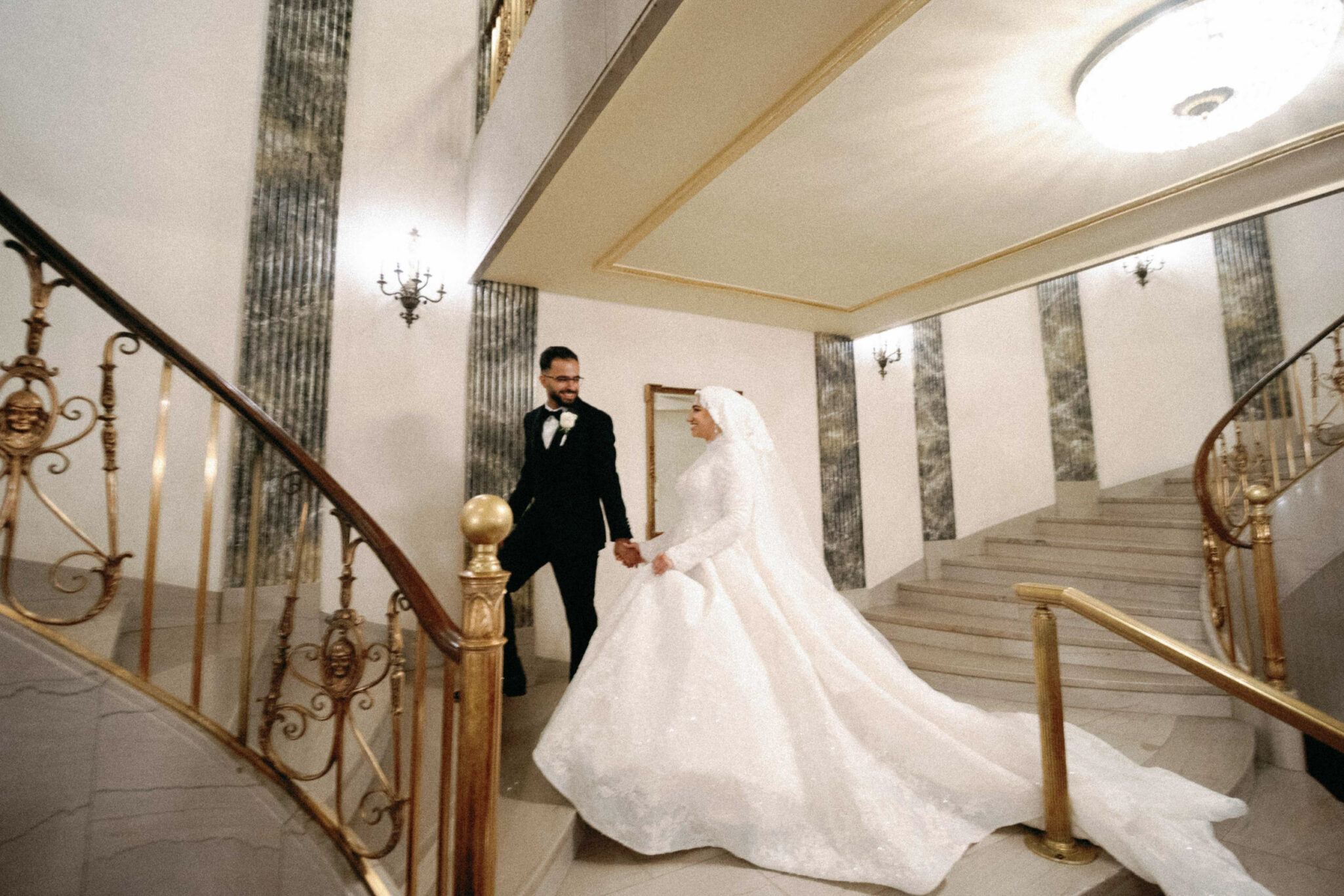 A bride in a white wedding gown and veil holds hands with a groom in a black tuxedo as they descend an elegant, curved staircase with gold railings—perfect for capturing timeless Arab wedding photography in Chicago’s grand, well-lit interiors.