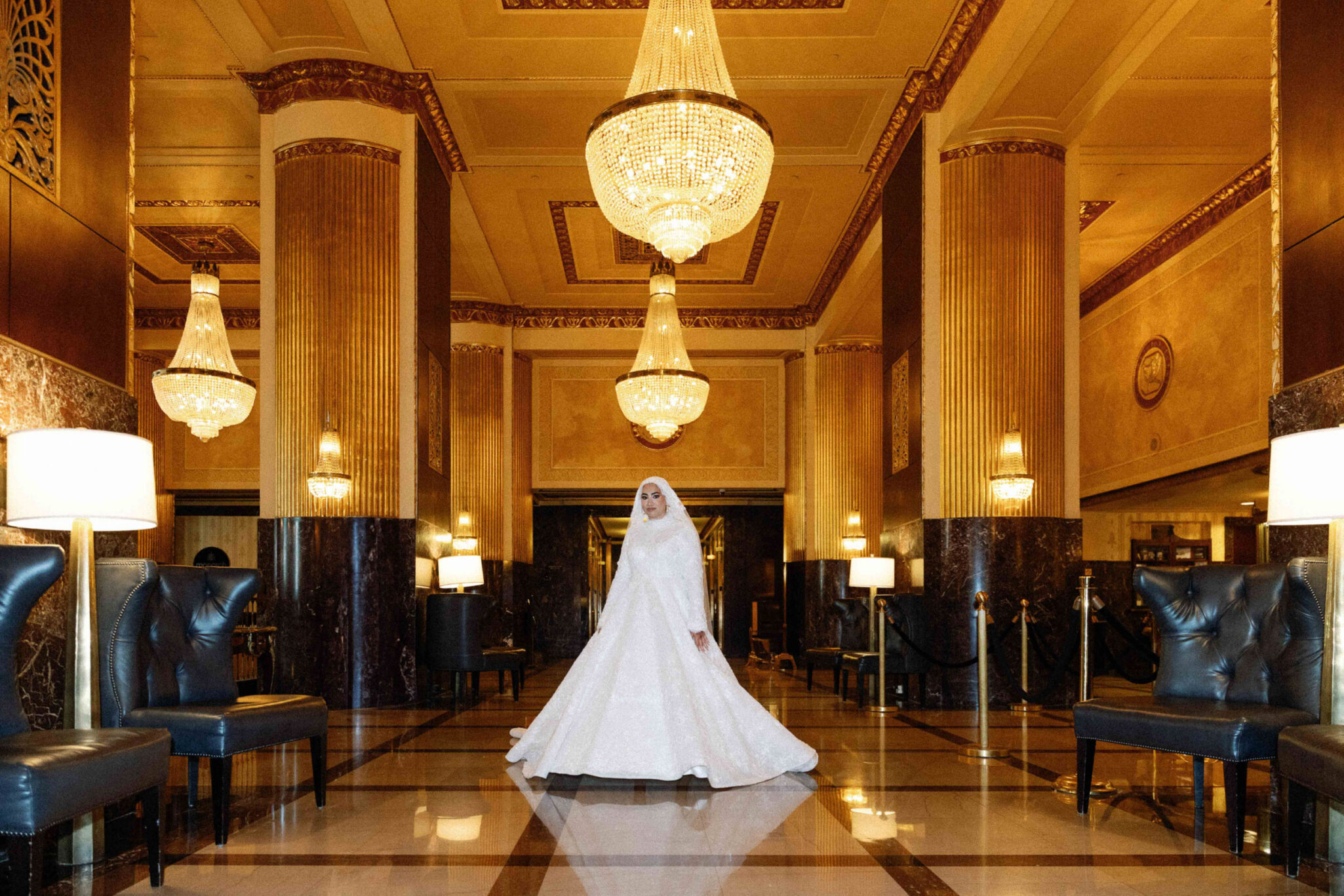 A bride in a white gown and veil stands in the center of an elegant, ornate hotel lobby—perfect for capturing Arab wedding photography in Chicago, with chandeliers, marble columns, and plush chairs creating a luxurious atmosphere.