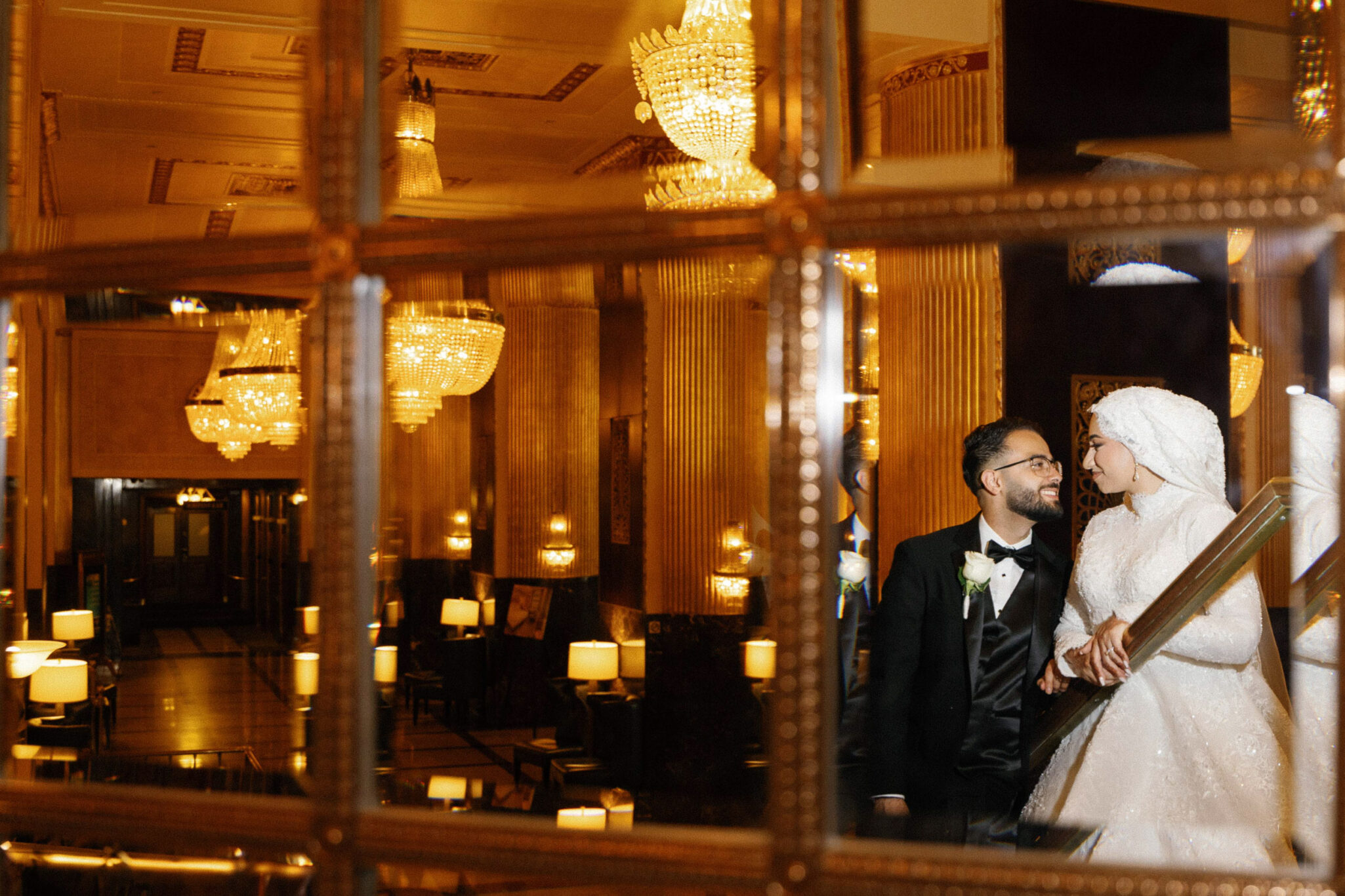 A bride in a white dress and hijab stands beside a groom in a black suit, smiling at each other in a grand, chandelier-lit hall, beautifully captured through expert Arab Wedding Photography with their reflection visible in a decorative mirror.