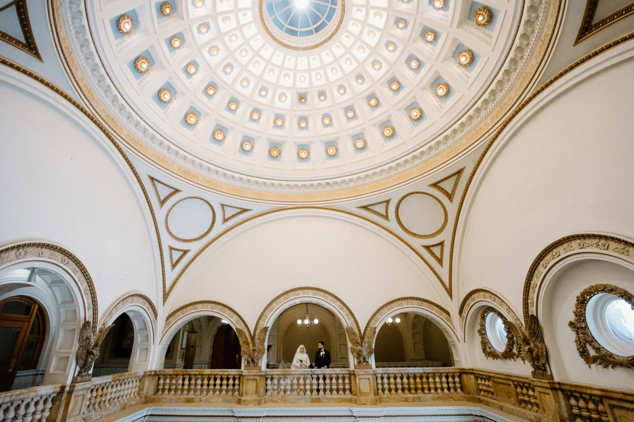 A bride and groom stand on an ornate balcony beneath a grand domed ceiling adorned with circular patterns and gold accents in an elegant, historic Chicago building—perfect for Arab Wedding Photography.