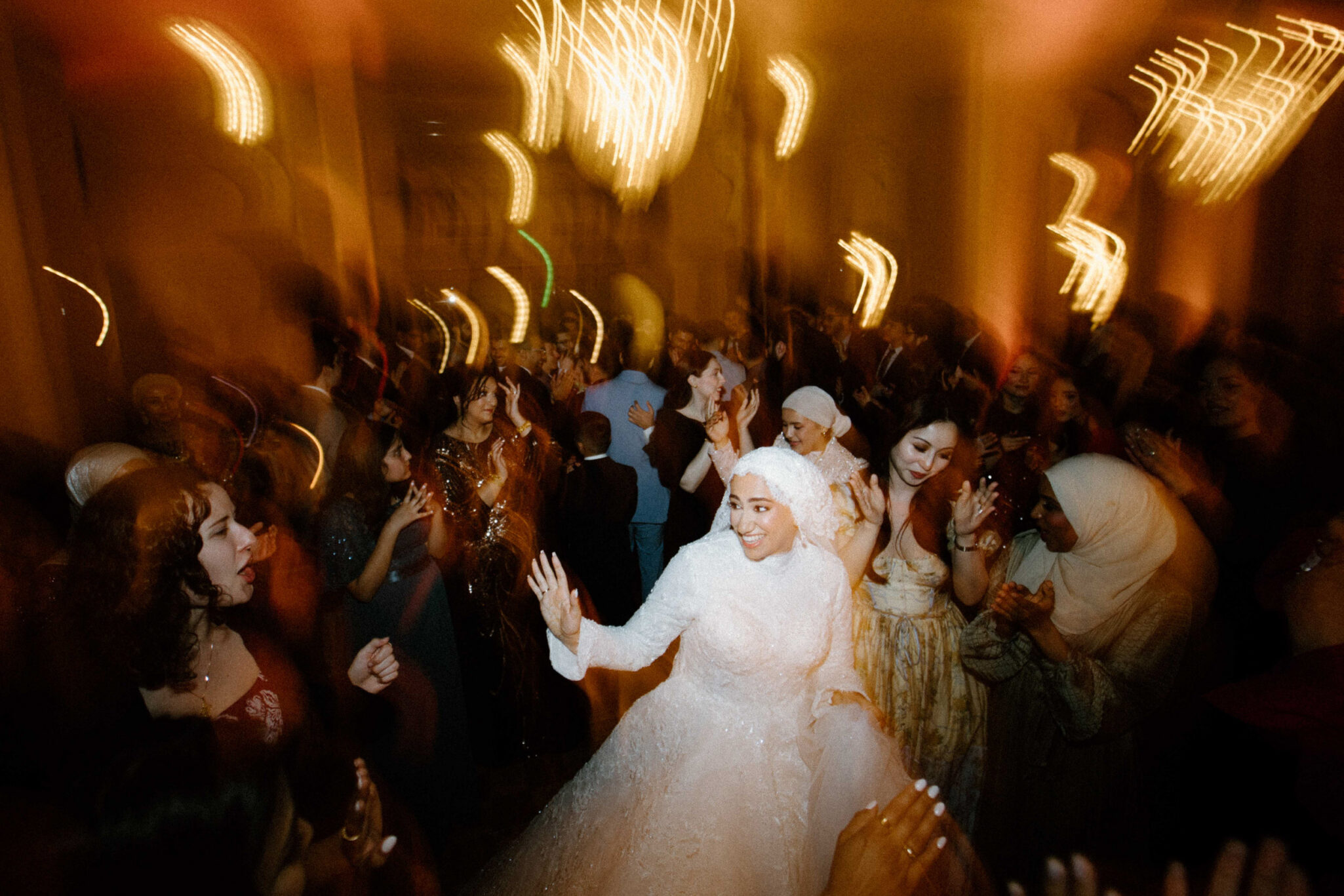 A bride in a white gown and hijab dances joyfully, surrounded by guests in elegant attire. Warm, swirling lights capture the festive energy of this multi-day Arab wedding photography and videography celebration.