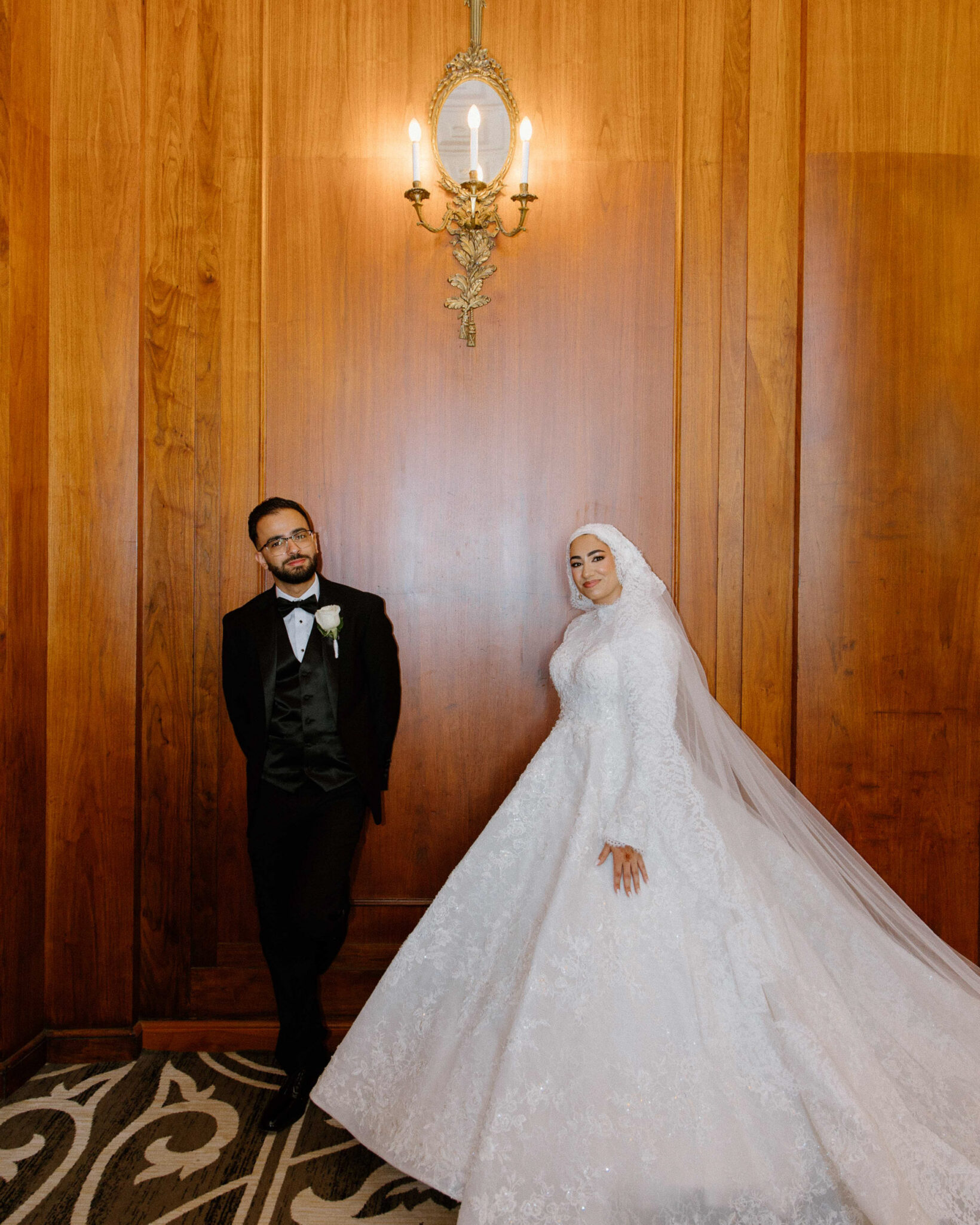 A bride in a white lace gown and hijab stands beside a groom in a black tuxedo, both posing against a wooden wall with an ornate sconce above them—a timeless moment captured in authentic Arab wedding photography in Chicago. The bride’s veil flows onto the patterned carpet.