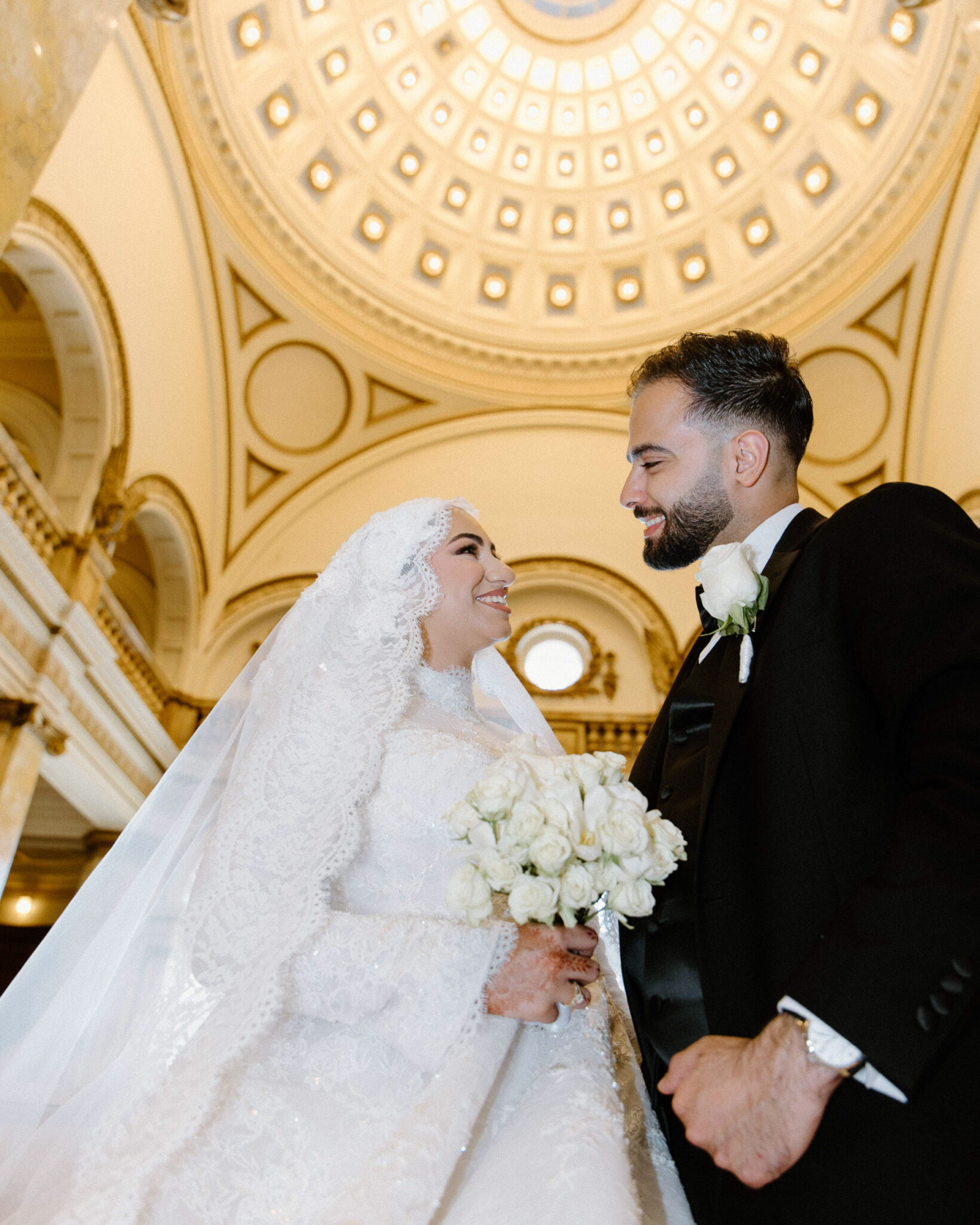 A bride in a white lace gown and veil holds a bouquet of white roses, smiling at the groom in a black suit beneath an ornate domed ceiling, beautifully captured by Chicago Arab wedding photography.