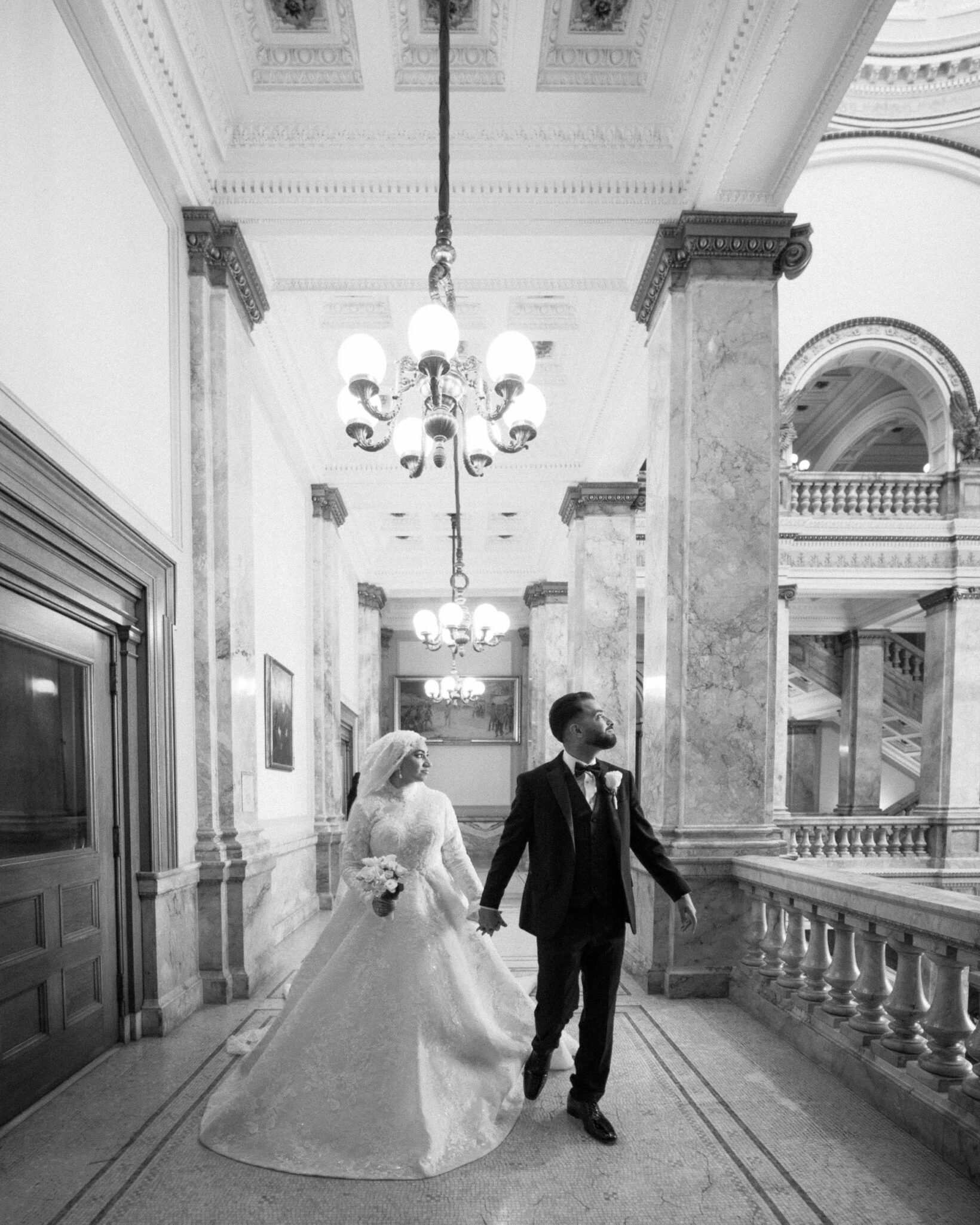 A bride and groom walk hand in hand down a grand marble hallway with ornate chandeliers and columns, dressed in formal wedding attire. The scene captures the elegance of Arab wedding photography in classic black and white.