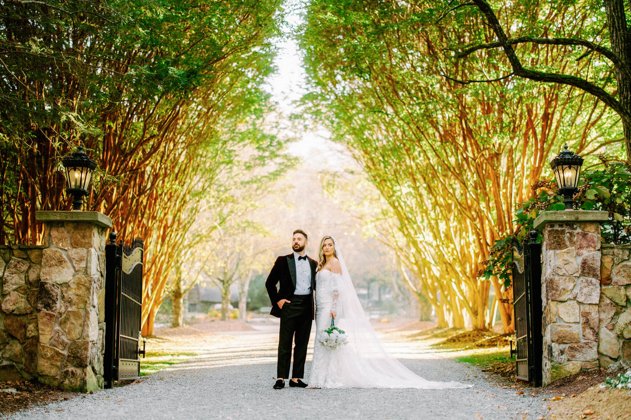 A bride and groom stand together on a tree-lined path at the entrance of a garden, dressed in wedding attire, with sunlight filtering through the branches above.
