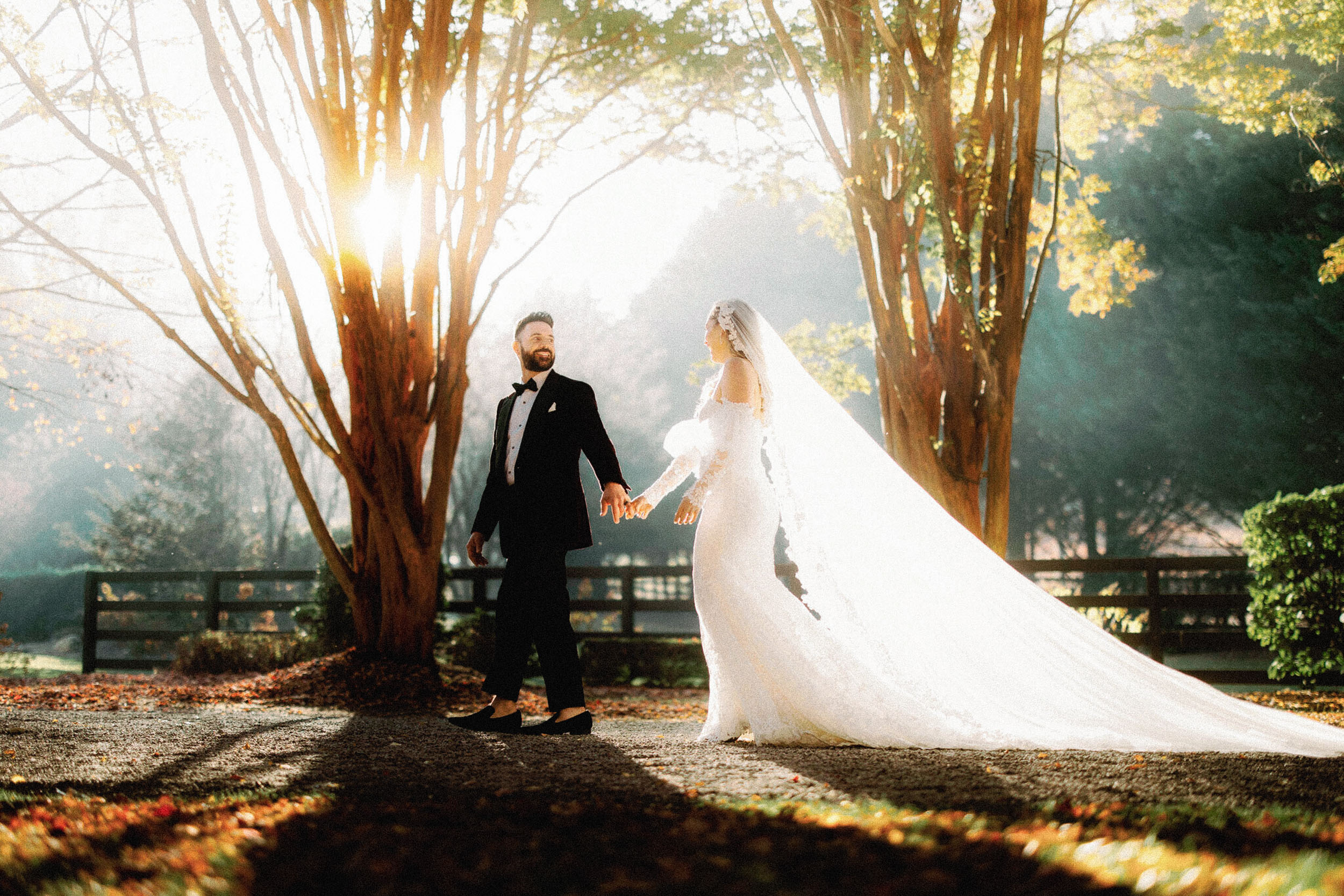 A bride in a long white gown and veil holds hands with a groom in a black suit as they walk outdoors in a sunlit, tree-lined Hawkesdene setting, capturing the magic of a full wedding weekend reunion style celebration.