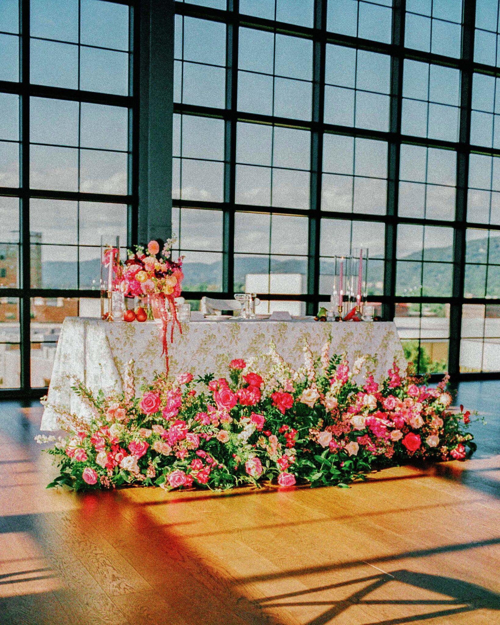 asheville wedding photographer A white table with floral arrangements and candles is set before large windows at the Biltmore Estate, surrounded by pink, red, and white blooms. Sunlight streams in, capturing a bright, elegant atmosphere perfect for film photography.