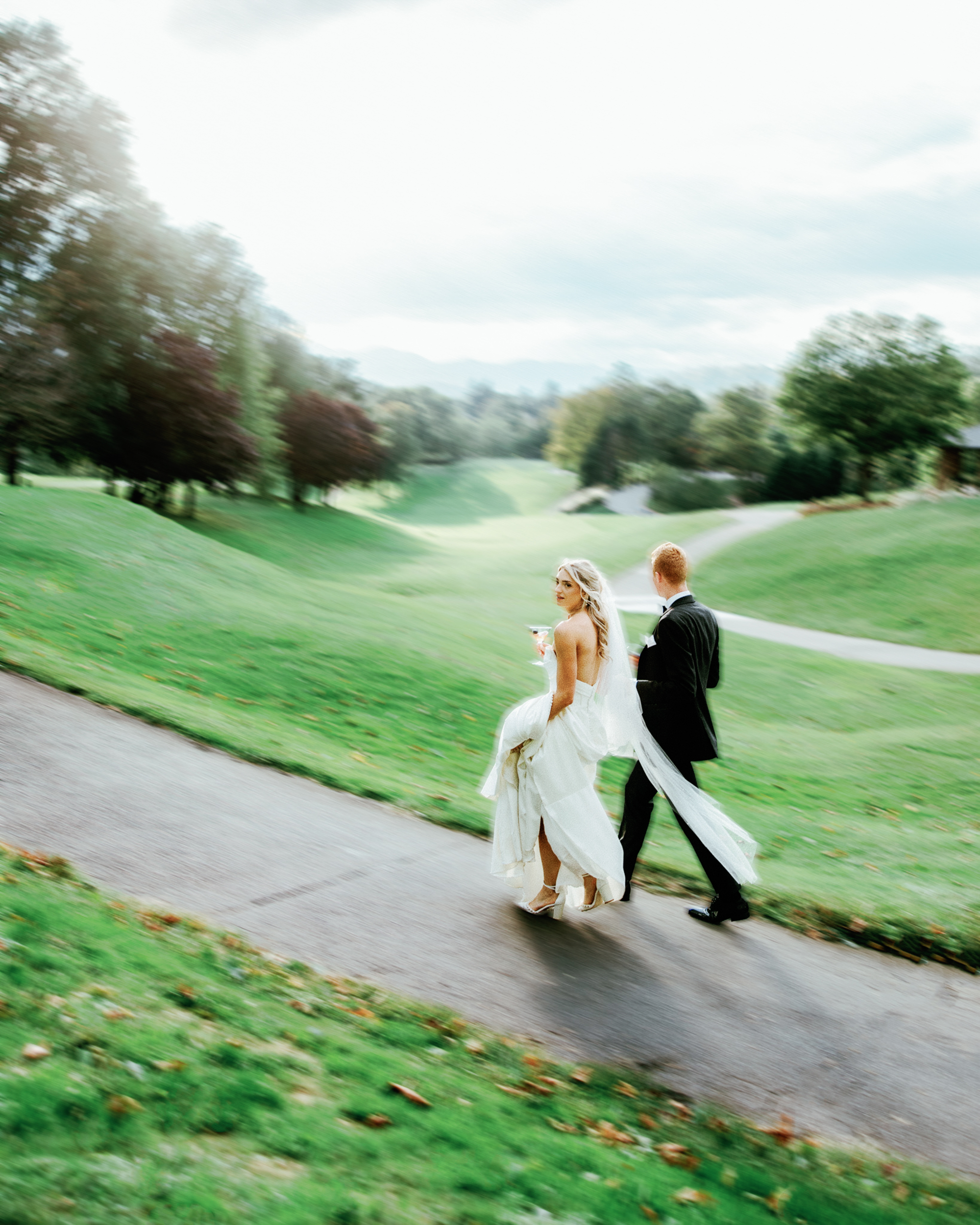 A bride in a white dress and veil walks with a groom in a black suit along a paved path, surrounded by greenery and trees, beautifully captured by The Omni Grove Park Inn wedding photographer.