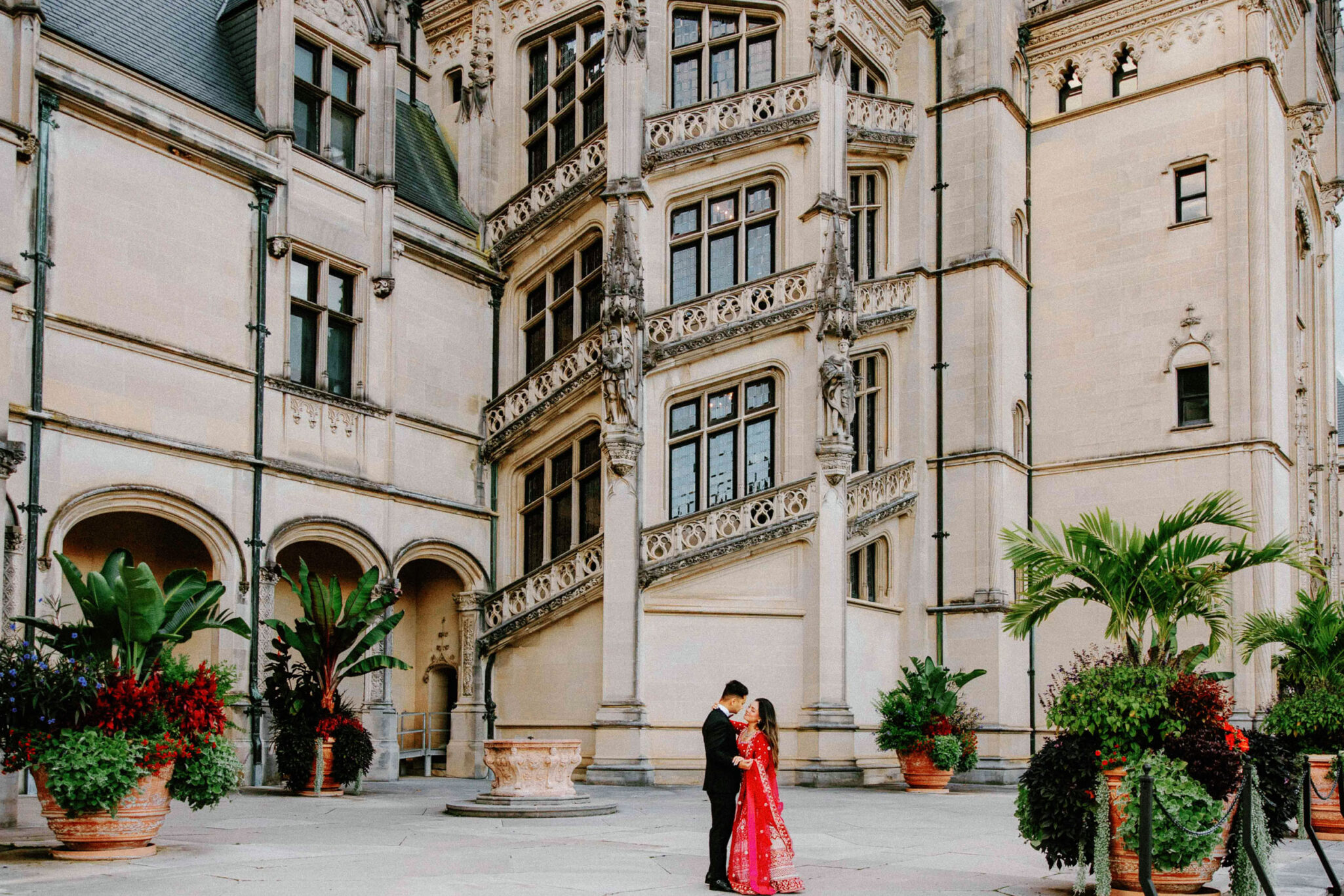 A couple, dressed in formal attire including a red dress, stands embracing in the courtyard of The Biltmore Estate, surrounded by its grand, ornate stone facade, large windows, elegant balconies, and lush potted plants.