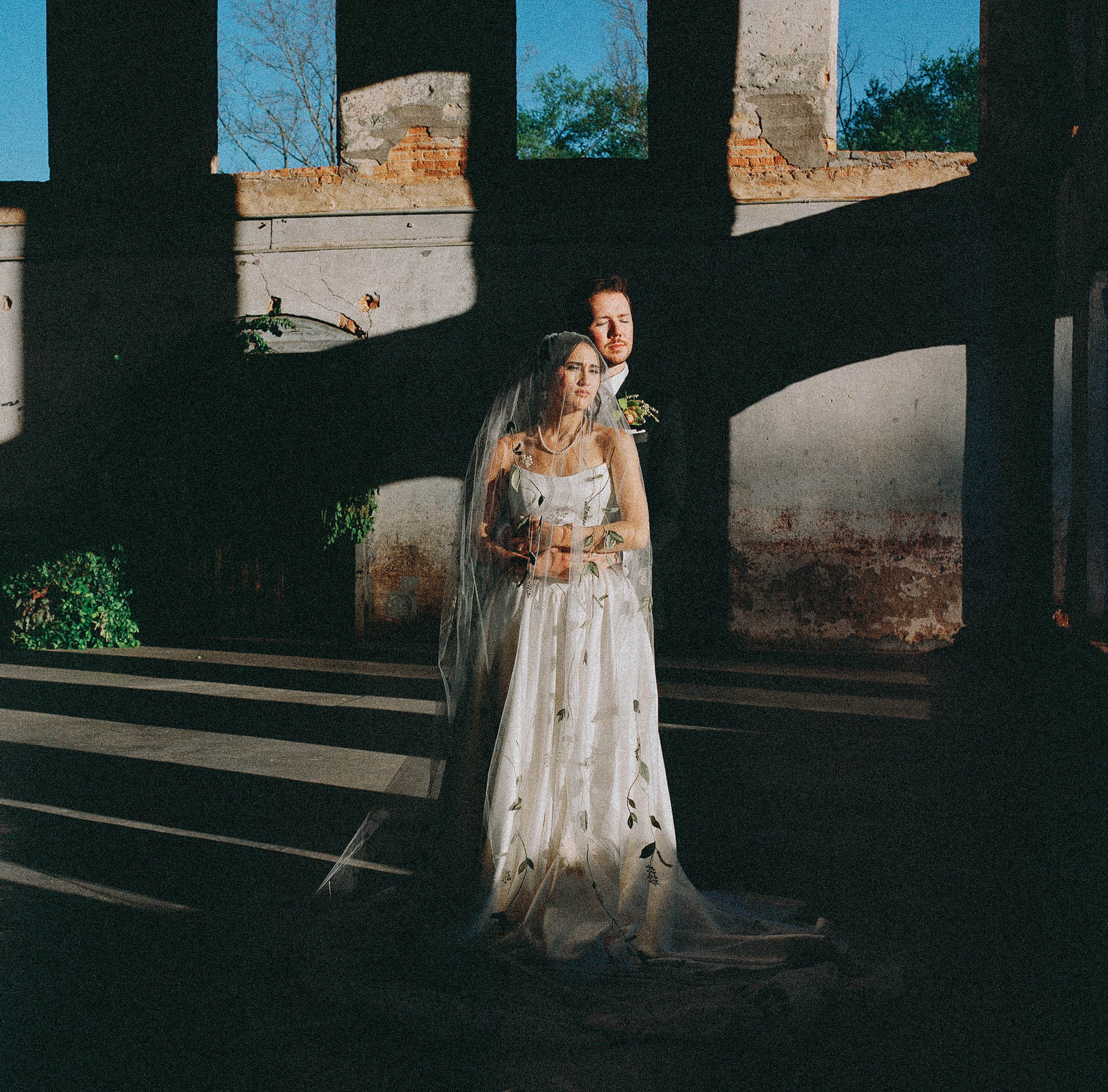 asheville wedding photographer A bride in a white dress and veil stands before a groom in a dark suit inside a sunlit, abandoned building, tall windows casting dramatic shadows—captured in timeless detail with 120 medium format film photography.