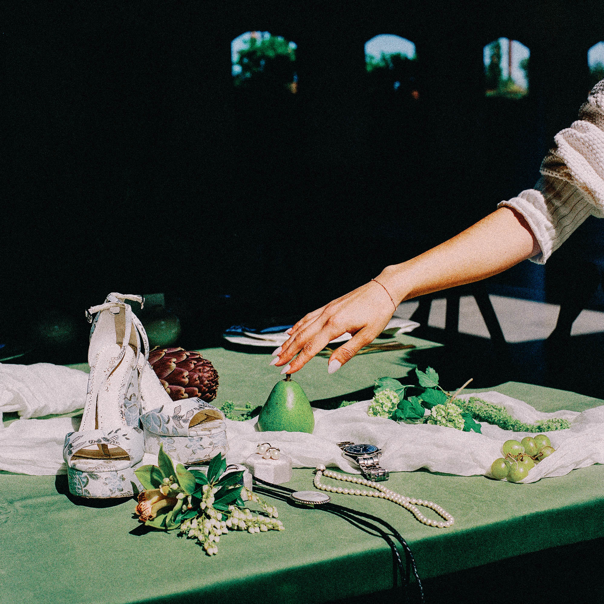 asheville wedding photographer A hand reaches toward a green pear on a table covered with green fabric, decorated with white shoes, jewelry, grapes, leafy greens, an artichoke, and a white cloth—all beautifully captured in 120 medium format film photography.