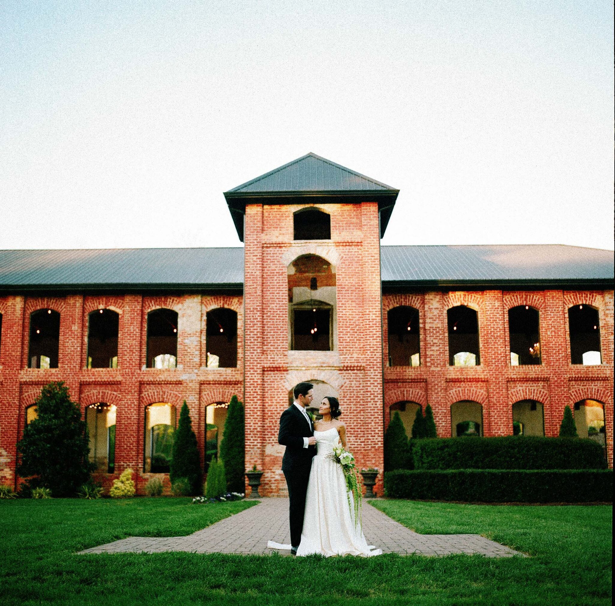 asheville wedding photographer A bride and groom stand together in front of a large, red-brick building with arched windows and a central tower, surrounded by green grass—captured in stunning detail on 120 medium format film photography under a clear sky.