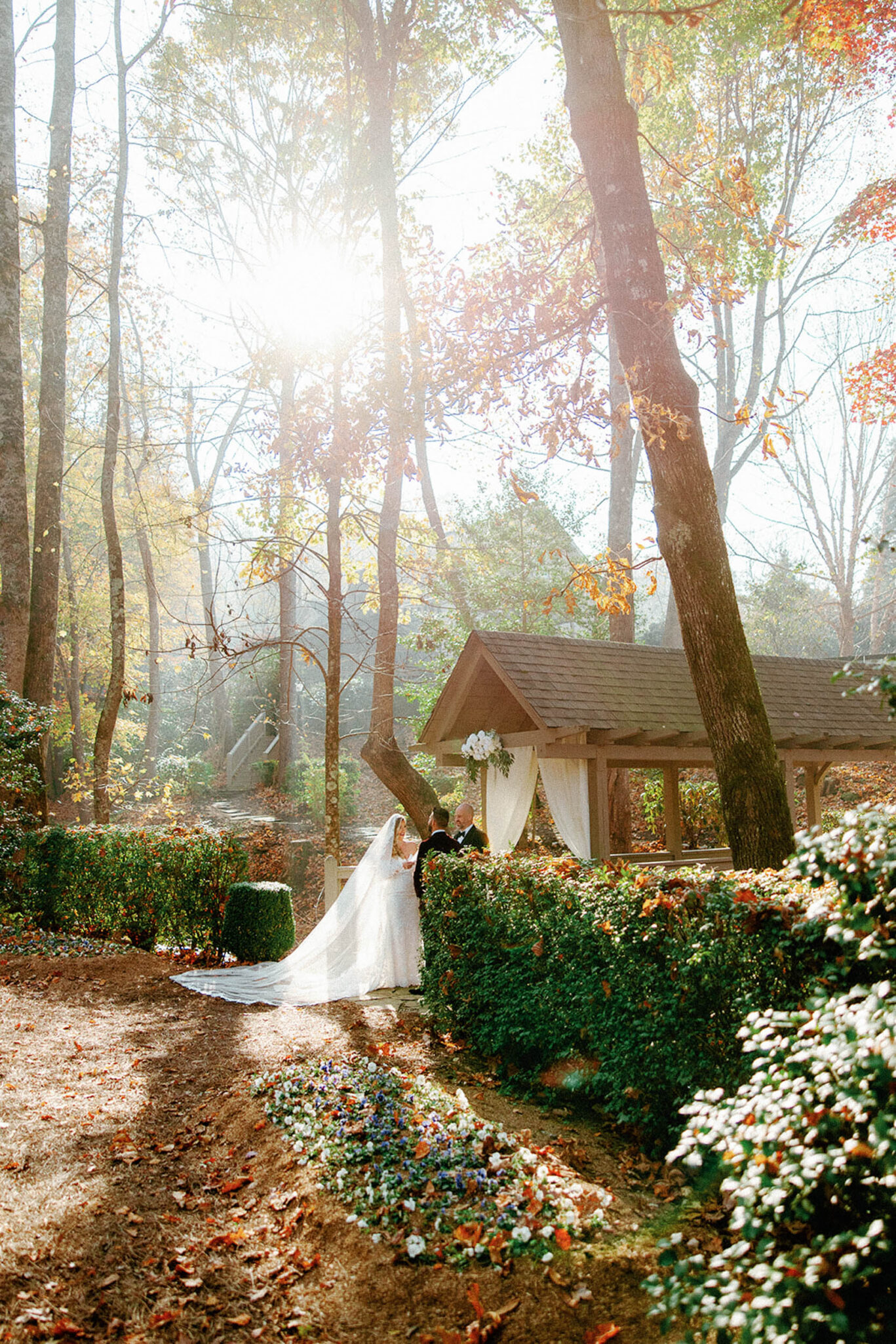 A bride and groom stand together in a sunlit garden, surrounded by tall trees and autumn foliage. The brides veil flows behind her as they pose near a flower-lined path and a small wooden pavilion draped with white fabric.