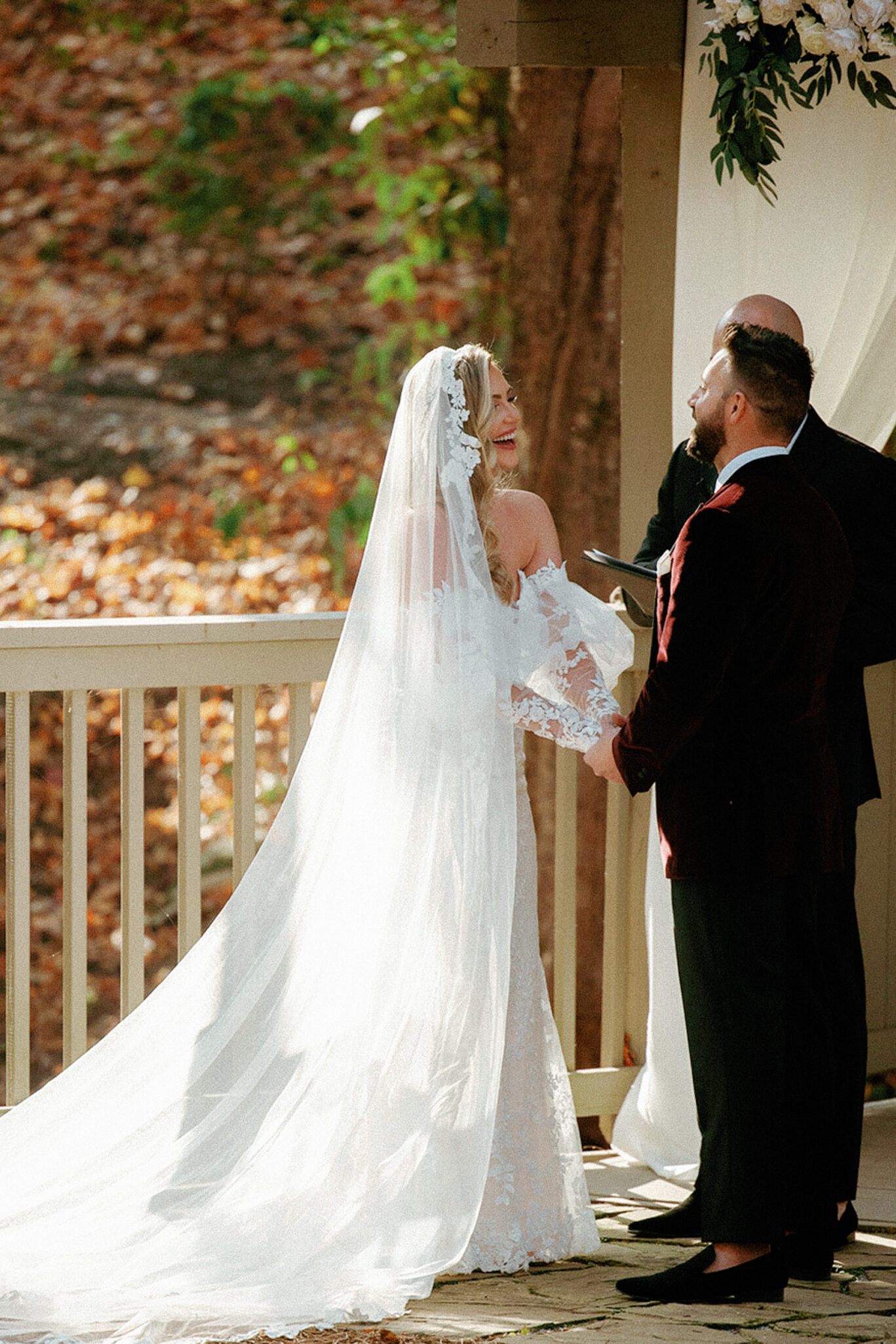 A bride in a long, white lace dress and veil smiles as she stands next to the groom during an outdoor wedding ceremony. They hold hands under a wooden gazebo decorated with flowers. Sunlight filters through autumn trees in the background.