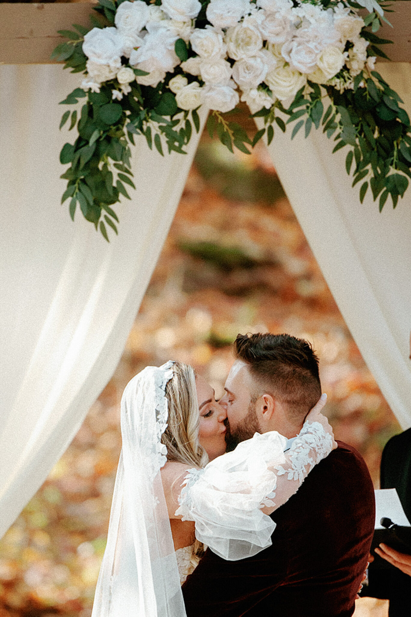 A bride and groom share a kiss under a floral arch with white flowers and greenery, framed by white drapes. The bride wears a lace veil and the groom has a dark beard and wears a suit.