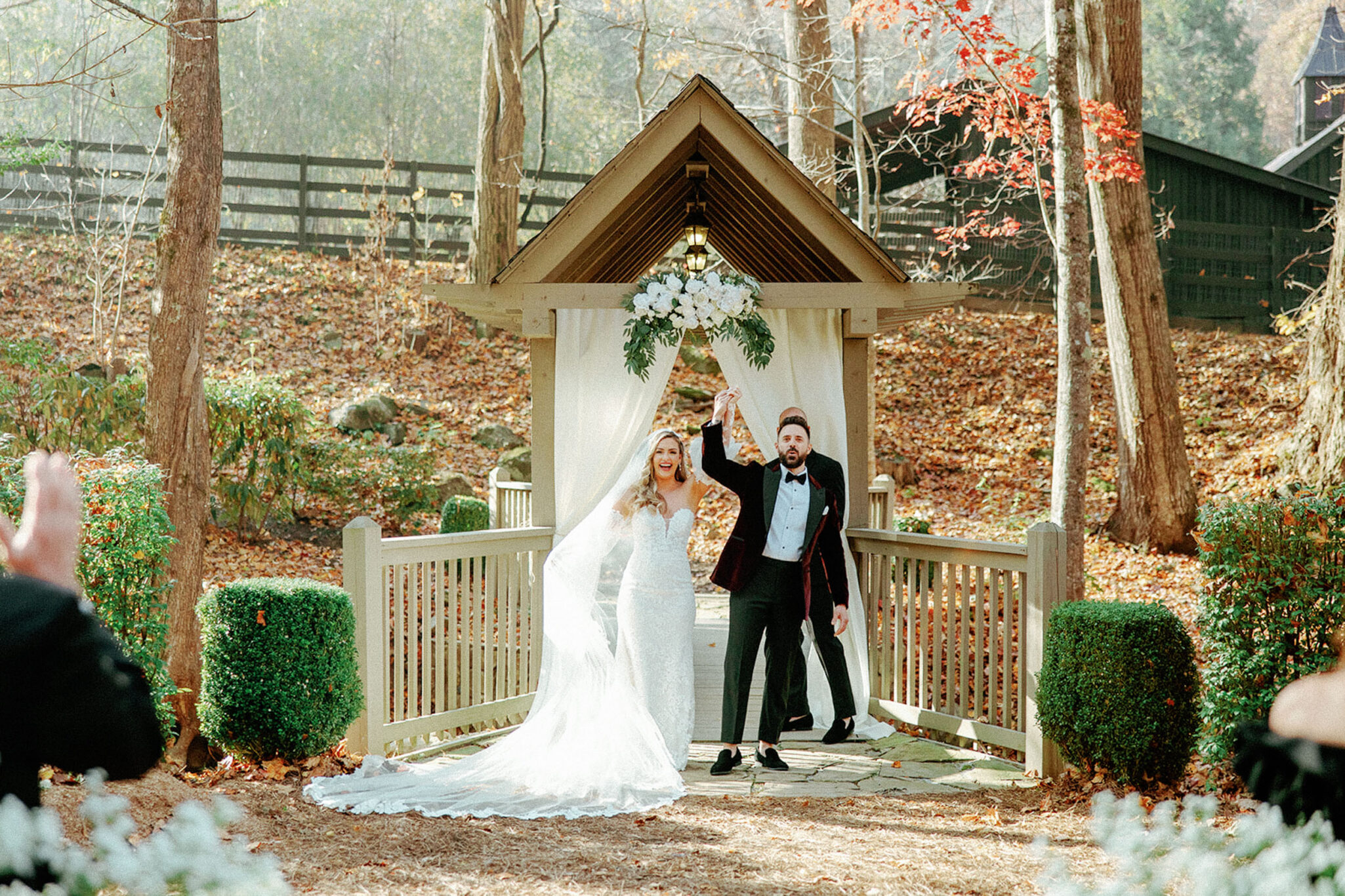 A bride and groom stand smiling under a small wooden pavilion outdoors, holding hands raised in celebration. The bride wears a long white dress with a veil; the groom wears a tuxedo. Autumn trees and guests are visible in the background.