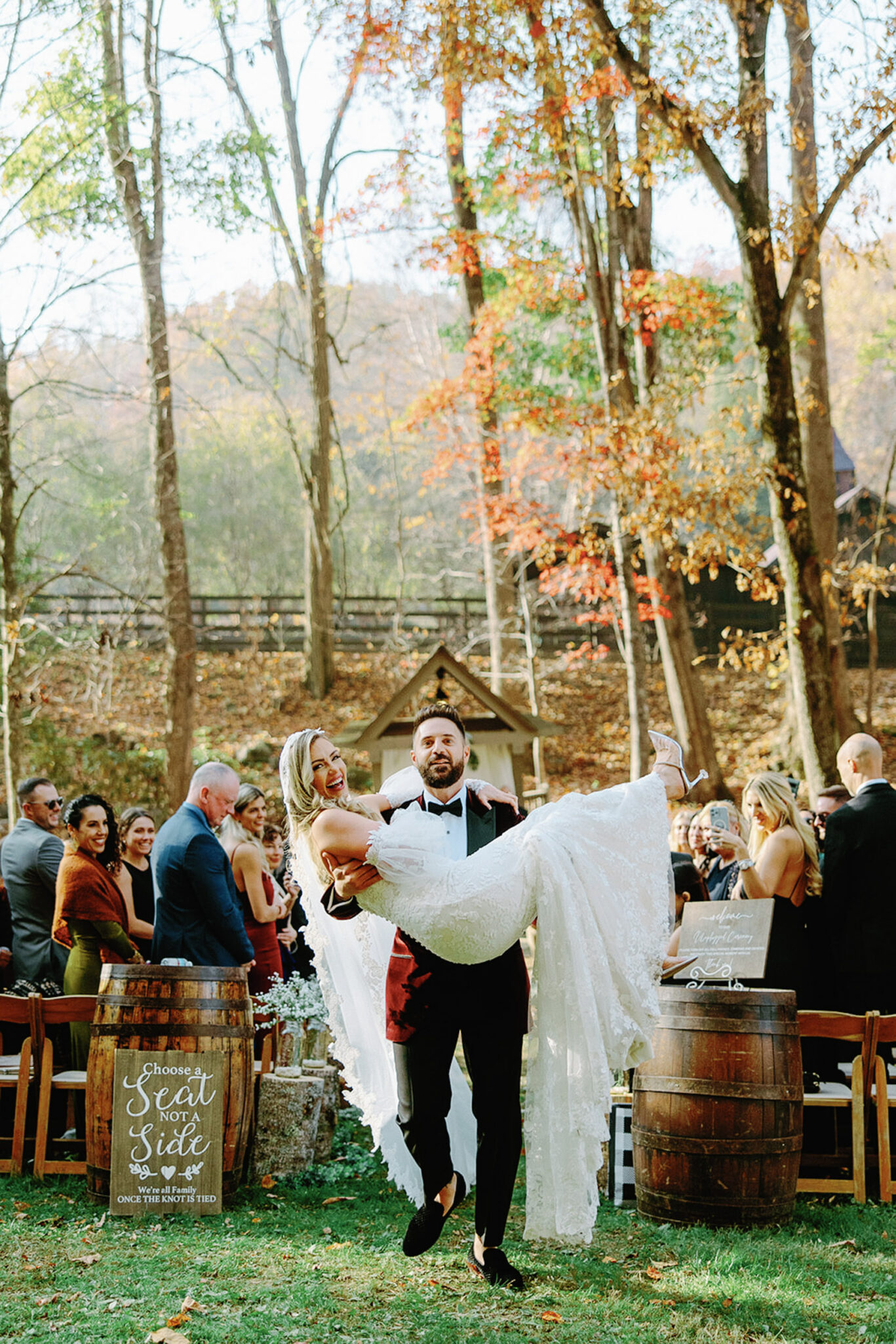A groom in a white jacket carries his smiling bride in a lace dress down an outdoor wedding aisle surrounded by guests, autumn trees, and wooden barrels. The scene is festive and joyful.