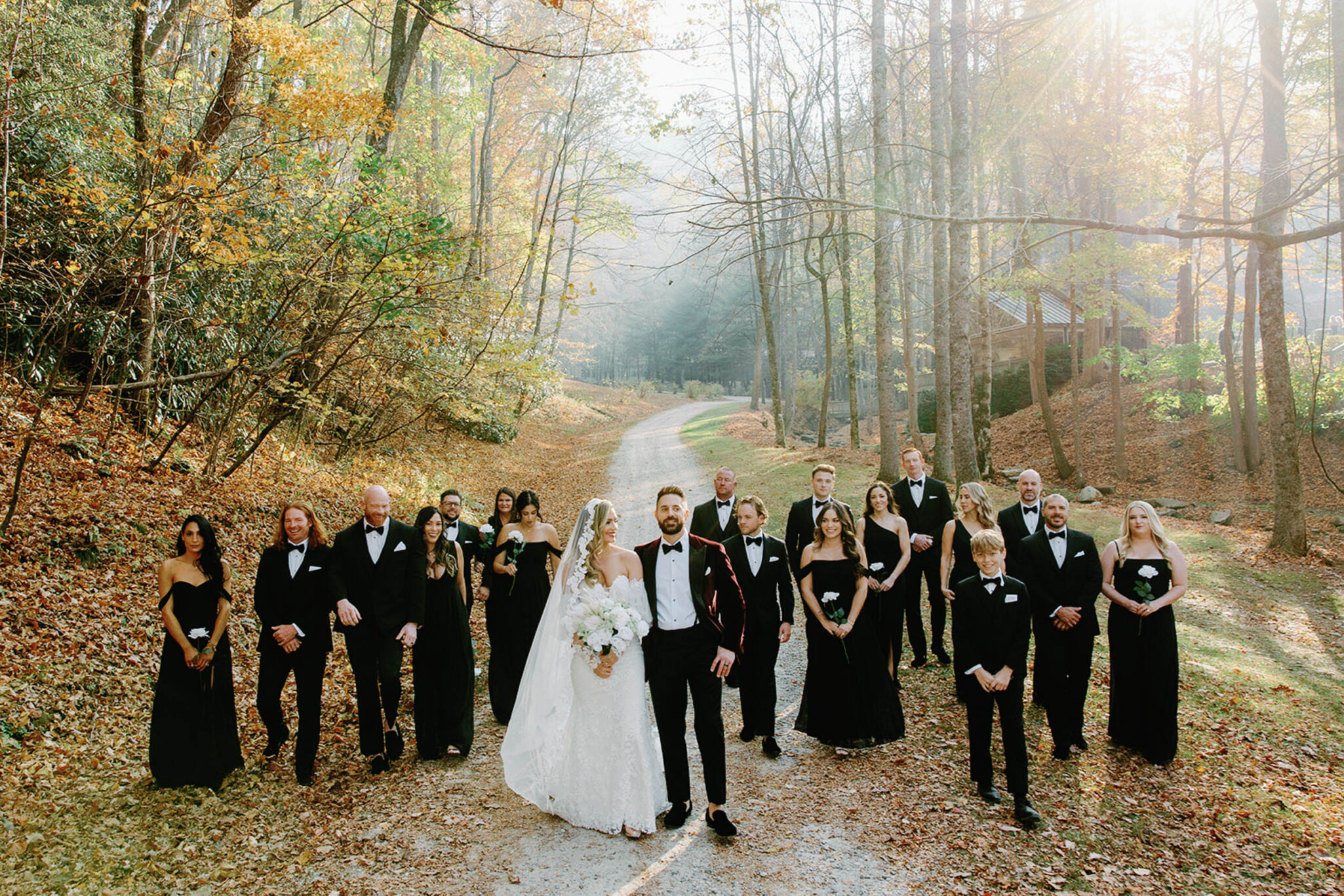 A bride and groom stand together on a wooded path, surrounded by their wedding party in black formal attire, with sunlight streaming through autumn trees and fallen leaves covering the ground.