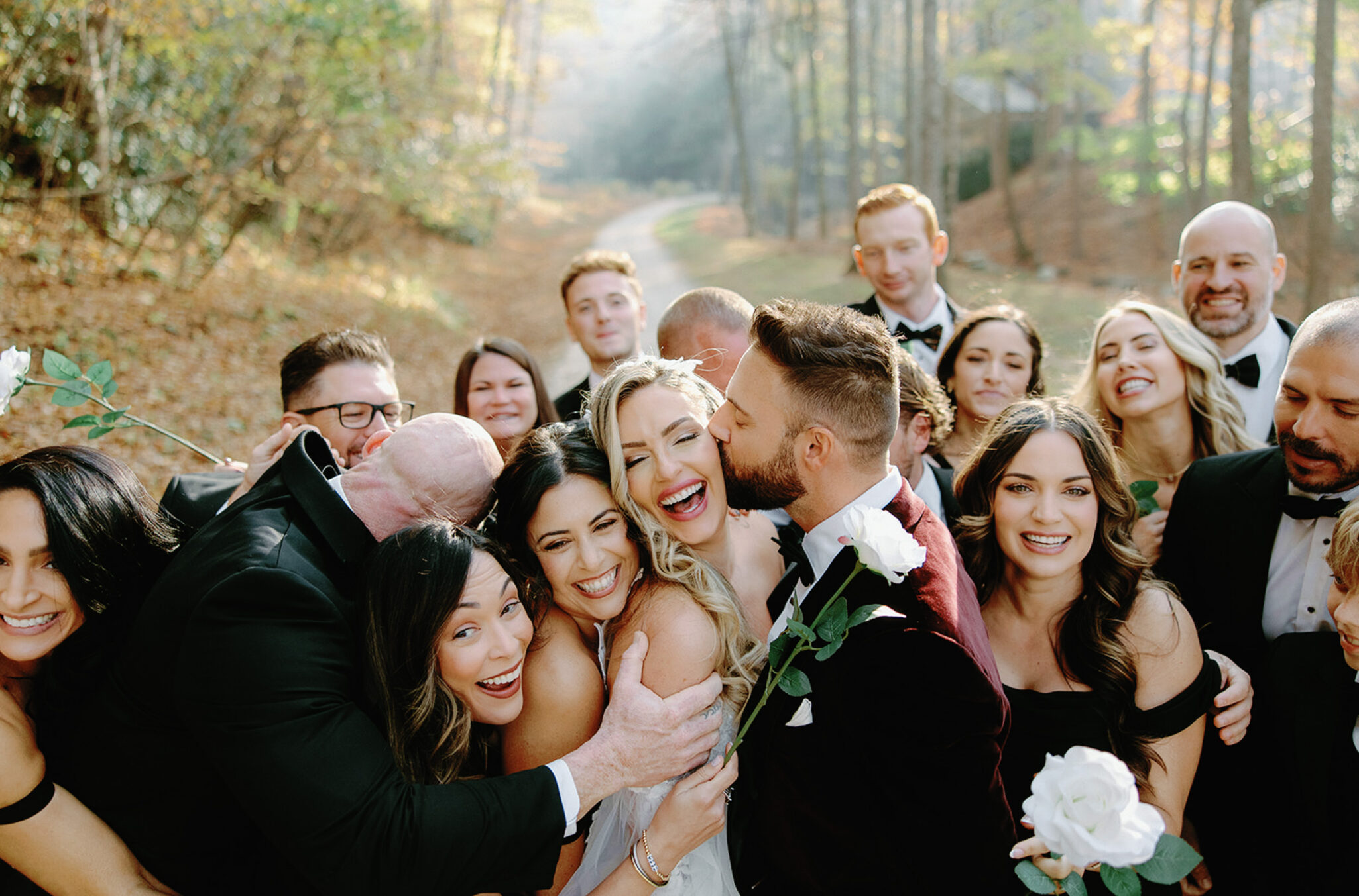 A joyful wedding party gathers outdoors, with the bride and groom smiling and embracing amid friends who hold white roses. Everyone appears happy and playful, surrounded by trees and sunlight filtering through.