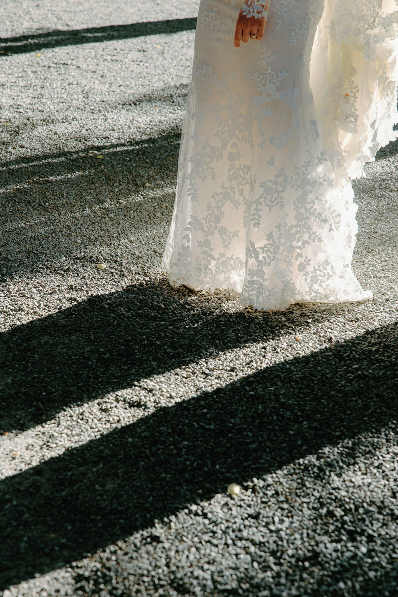 A person in a white lace wedding dress stands on gravel, with sunlight casting long shadows. Only the lower half of the dress and the persons hand are visible.