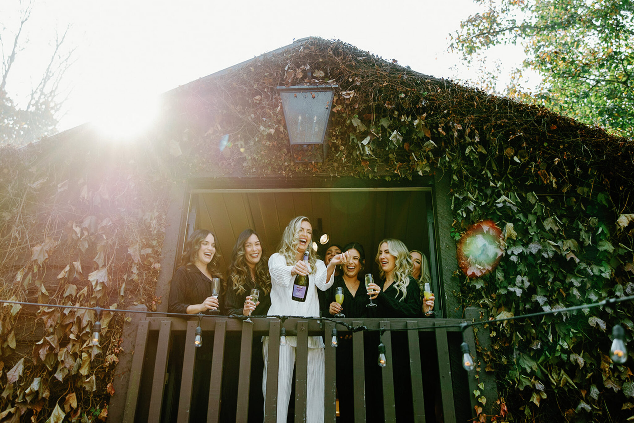 A group of women stand on a vine-covered balcony, laughing and cheering as one pops a bottle of champagne. Sunlight streams in from the left, creating a warm, celebratory atmosphere.