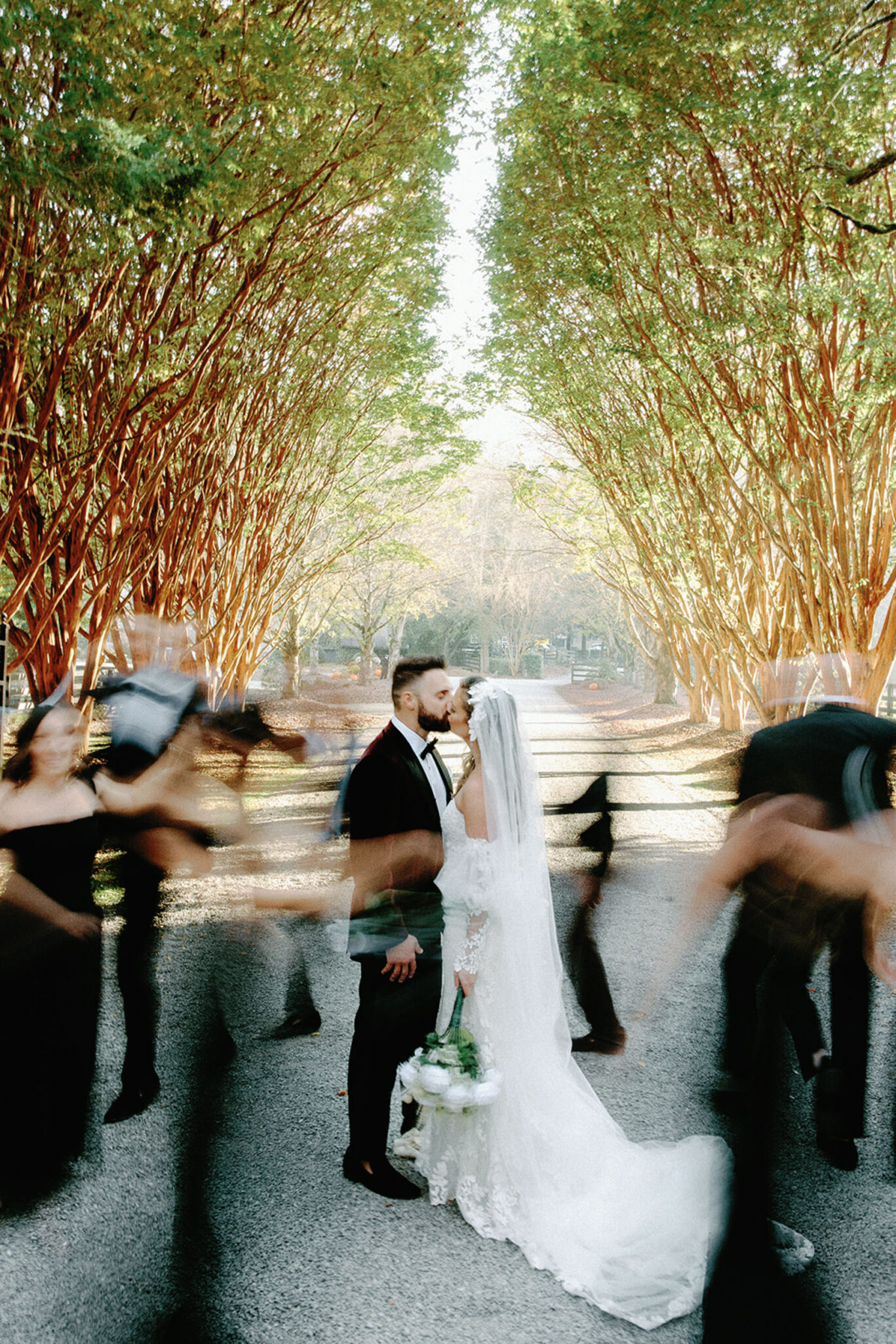 A bride and groom stand facing each other on a tree-lined path, sharing a quiet moment. Blurred figures in black move around them, highlighting the couple’s stillness and intimacy. Sunlight filters through the green canopy above.