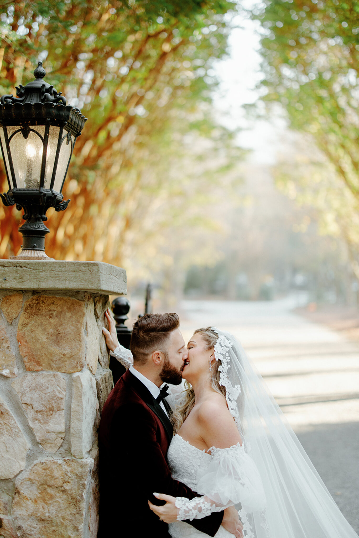 A bride and groom share a romantic kiss beside a stone wall and vintage lamp, surrounded by trees and bathed in soft, golden sunlight. The bride wears an off-shoulder lace dress and veil, while the groom wears a dark suit.