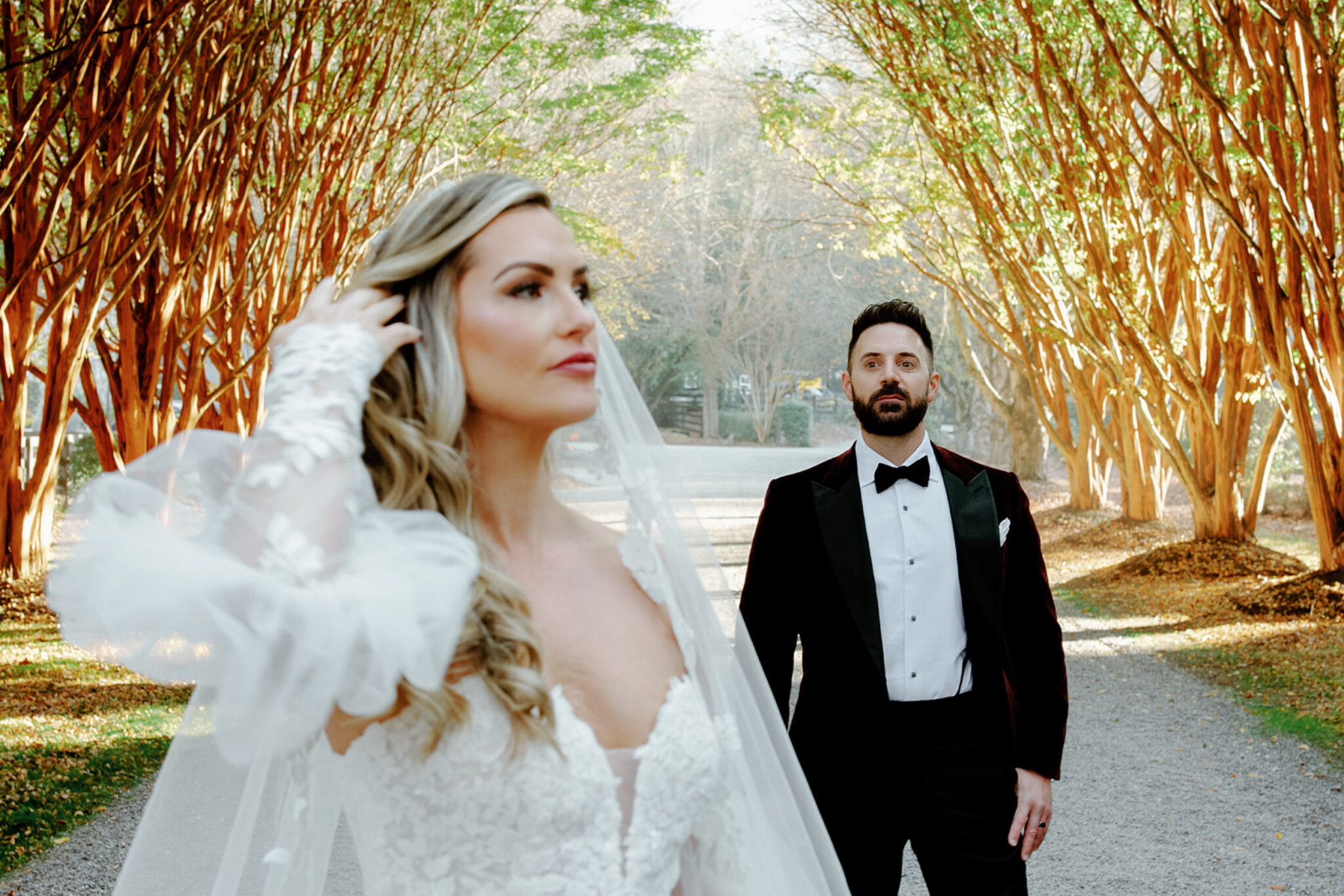 A bride in a white lace wedding dress stands in the foreground, adjusting her hair, while a groom in a black tuxedo stands behind her on a tree-lined path with sunlight streaming through the branches.