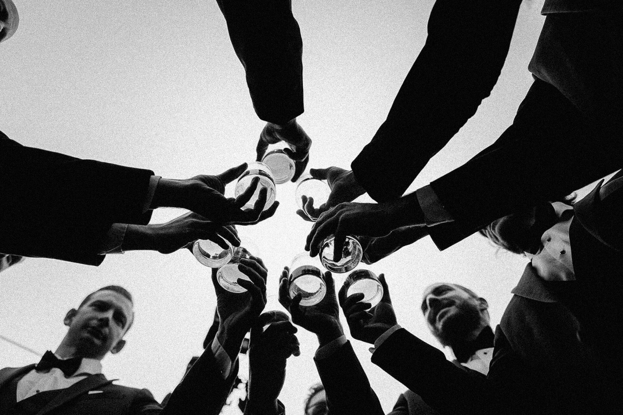 A group of people in formal attire raise glasses together in a celebratory toast, viewed from below against a bright sky.