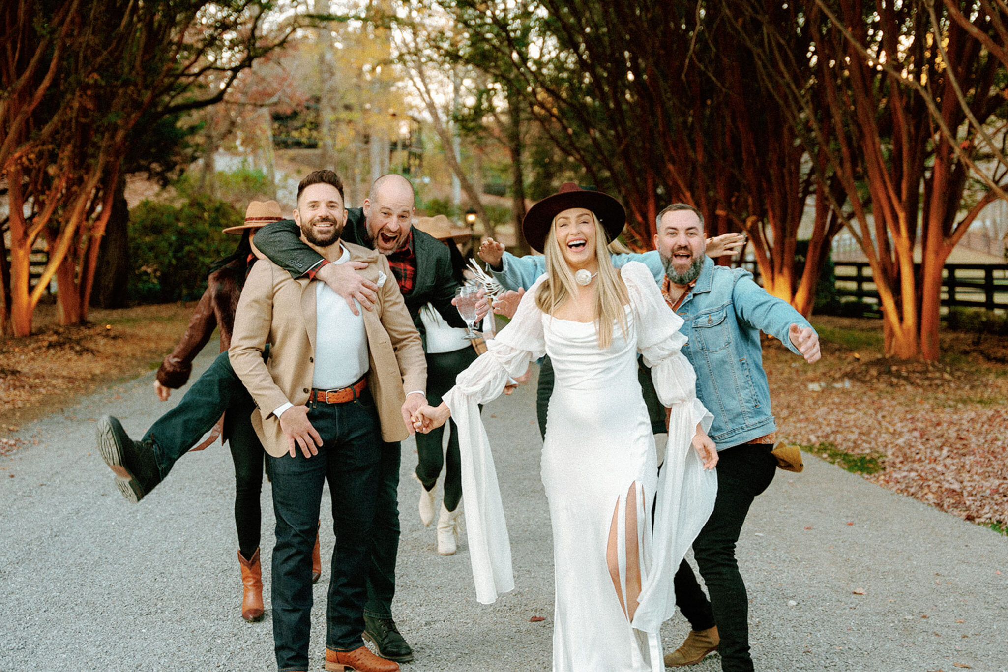 A bride in a white dress and hat joyfully leads a group of men down a tree-lined path at Hawkesdene; everyone is smiling, laughing, and playful, capturing the full wedding weekend’s reunion-style celebration.