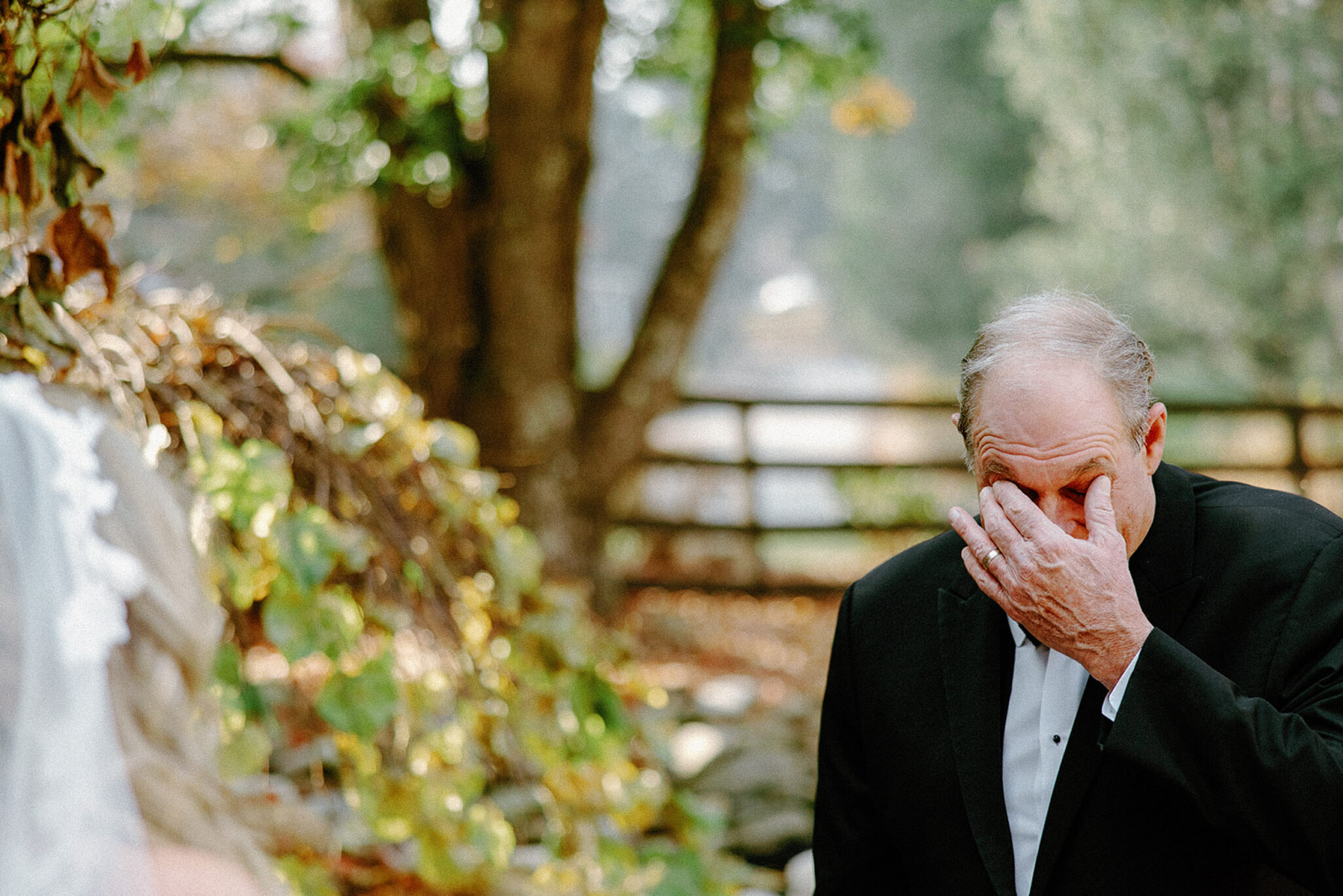 A man in a tuxedo wipes his eyes, appearing emotional, while standing outdoors near greenery and a wooden fence. The blurred figure of a person in a white veil is visible on the left.