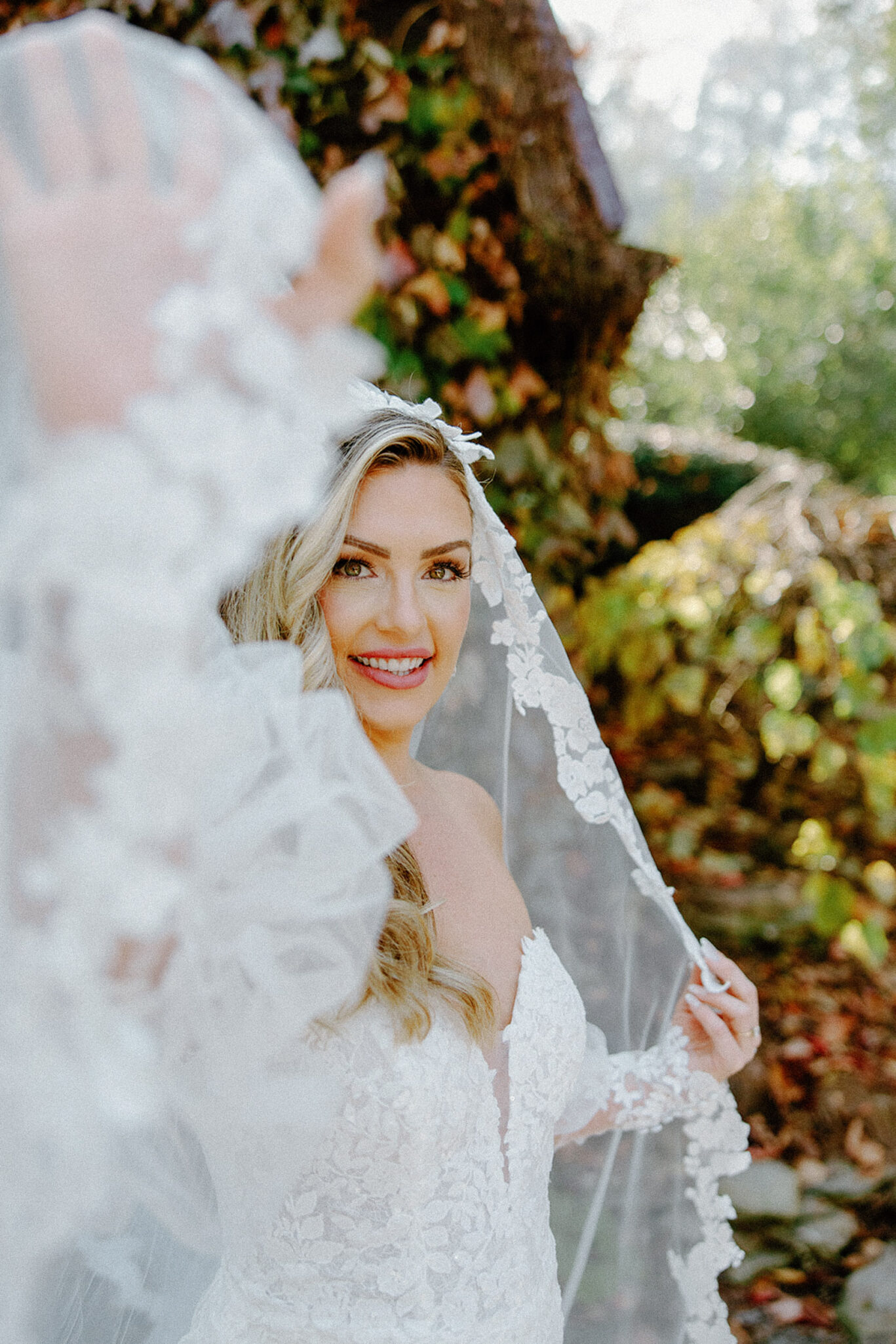 A smiling bride in a white lace wedding dress holds up her veil outdoors, surrounded by greenery and autumn leaves. Sunlight highlights her wavy hair and joyful expression.