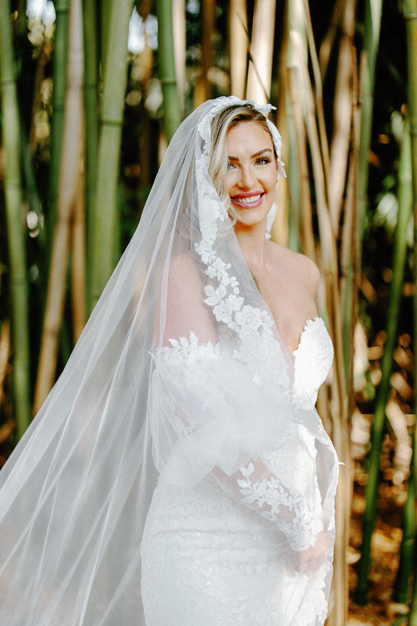 A smiling bride in a white lace wedding dress and veil stands in front of tall green bamboo stalks, holding the veil around her shoulders.